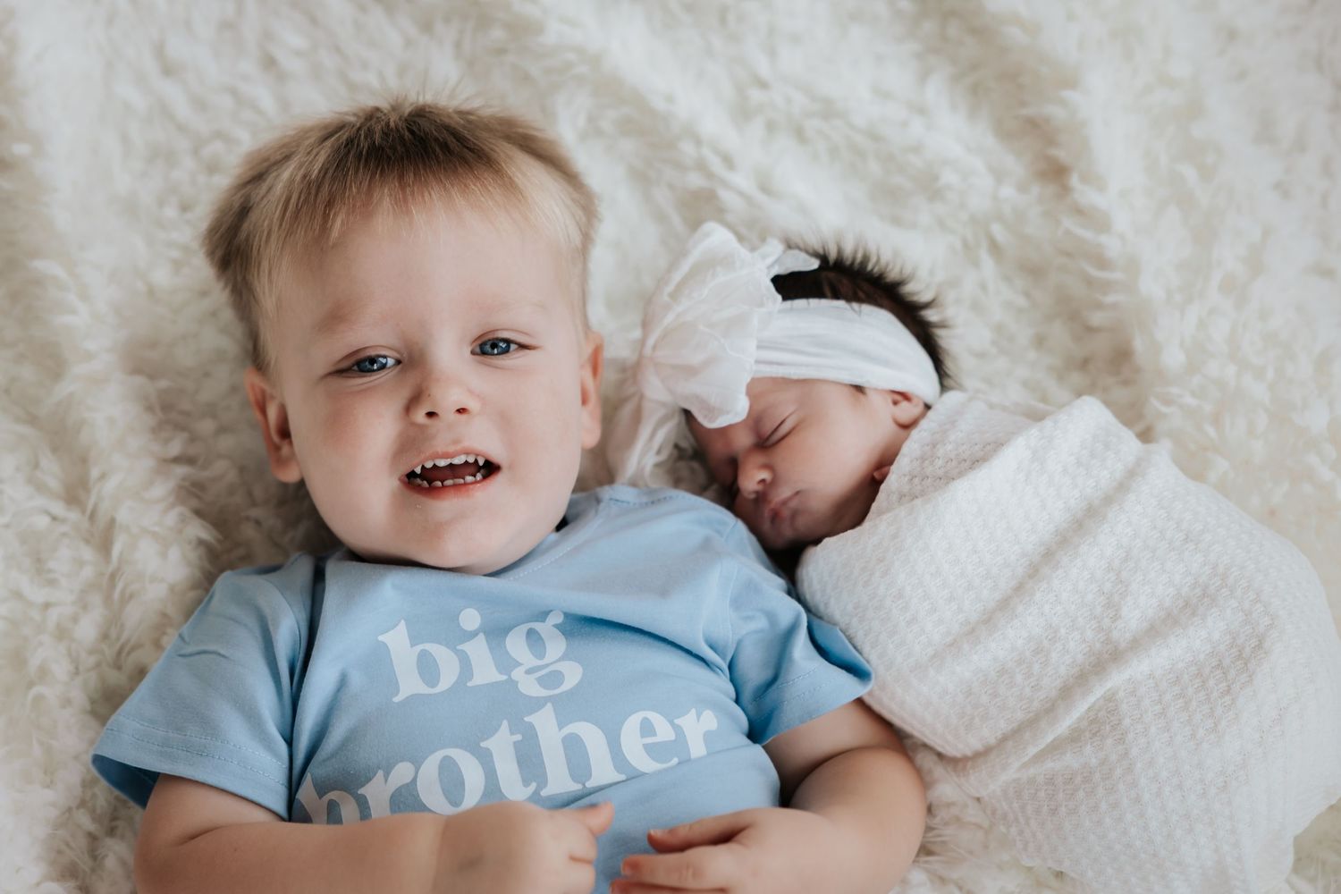 Toddler boy in a blue big brother t-shirt lies beside his sleeping swaddled newborn sister on a soft cream blanket.
