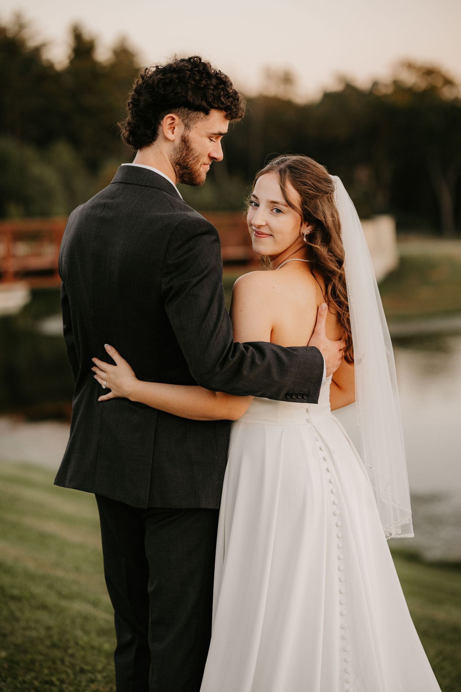 Smiling bride glances over her shoulder as her bearded groom holds her close on a grassy lakeside at sunset.