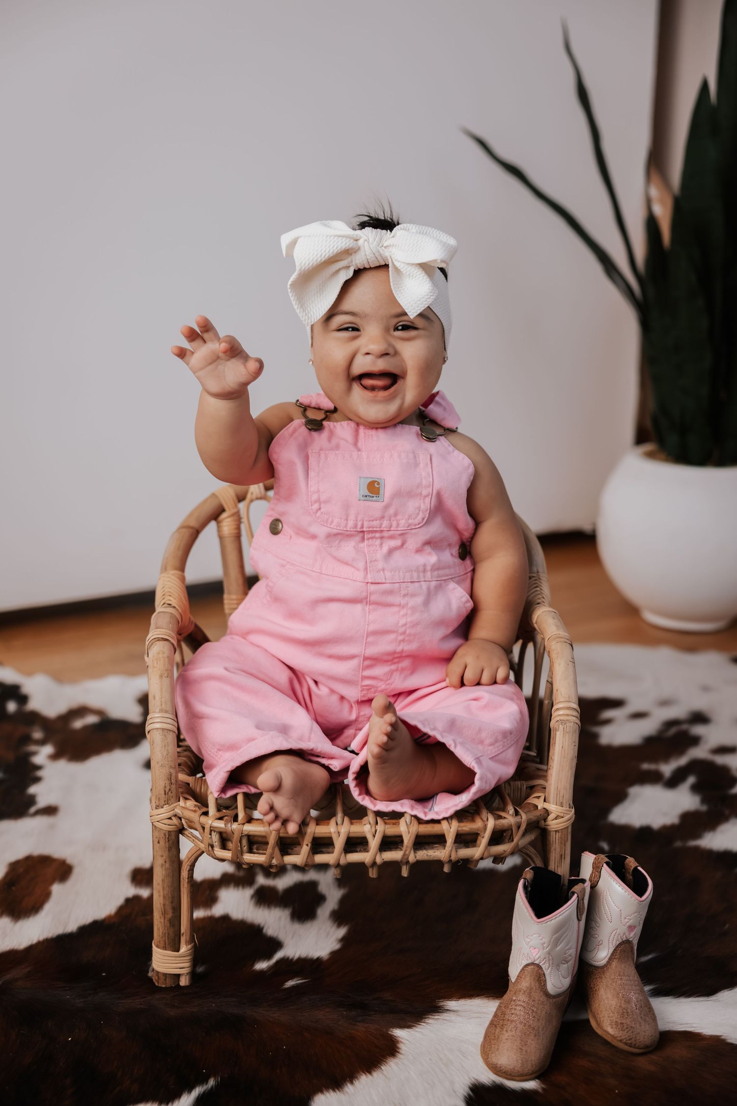 Smiling baby in pink overalls and a white bow waves from a rattan chair on a cowhide rug with small cowboy boots nearby.
