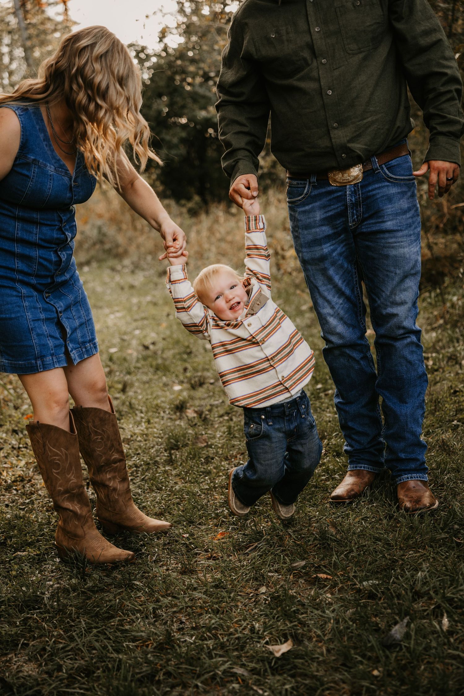 Parents swing their giggling toddler by the hands in a grassy outdoor setting, wearing denim, boots, and western style clothing.