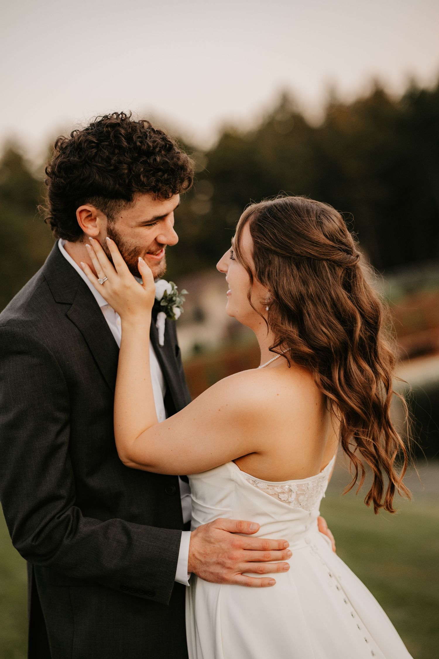 Smiling groom in a dark suit looks at his bride in a white dress as she gently touches his face during sunset wedding portraits.