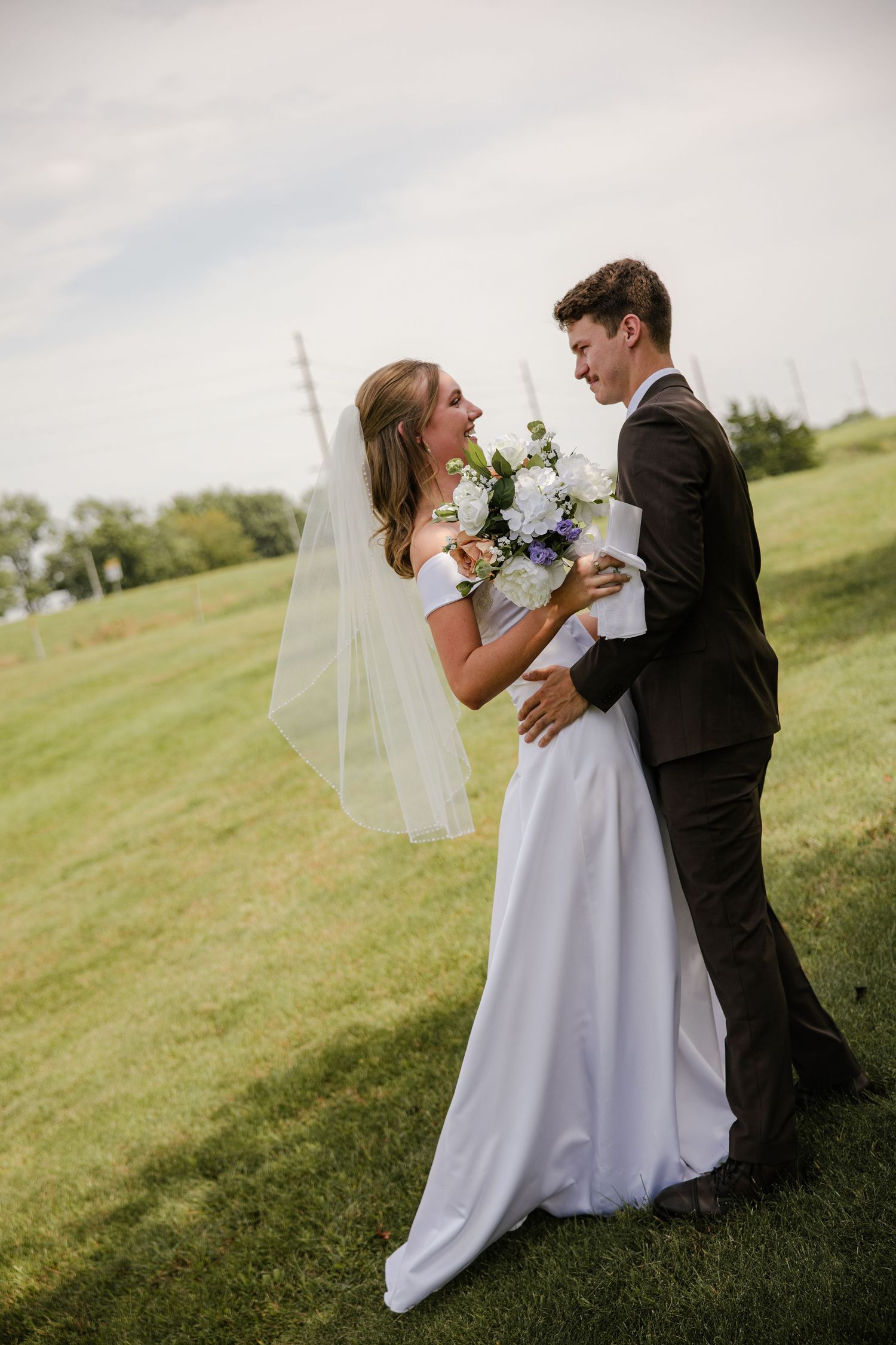 Bride in a white gown and veil smiles at her groom while holding a bouquet in a sunny grassy field.