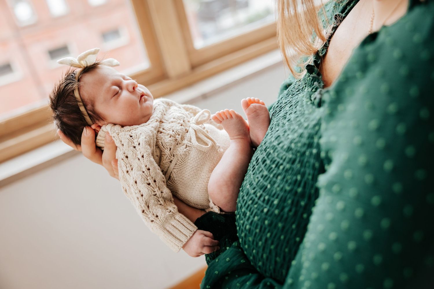 Close-up of a sleeping newborn with bare feet and a bow headband held gently in a mother's arms by a window.