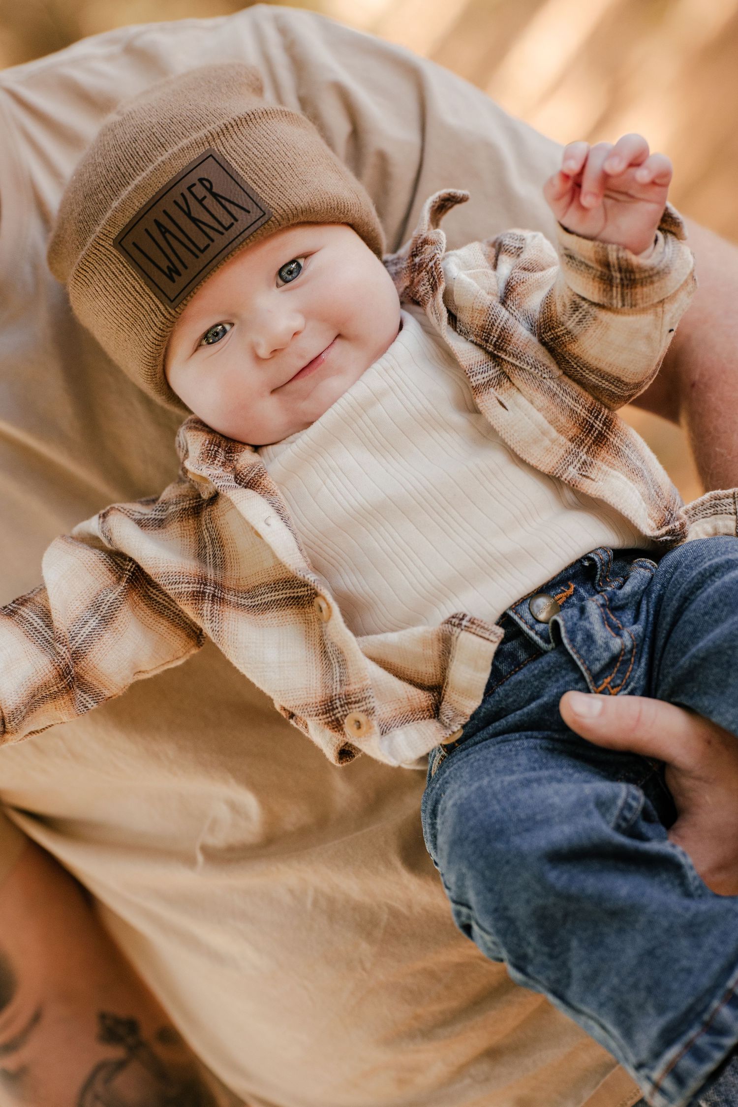 Cheerful baby in a WALKER beanie, cream top, plaid flannel and jeans rests in a parent's arms with one hand raised.