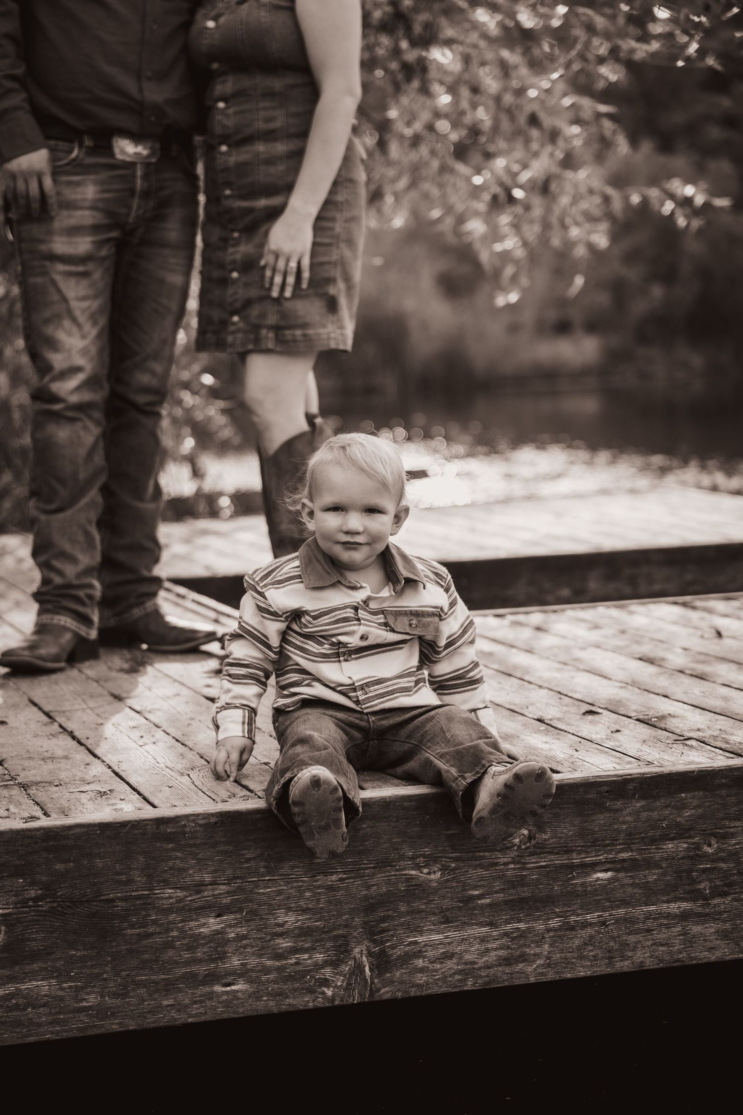 Black and white photo of a toddler seated on a wooden dock with his parents standing behind him in jeans and boots.