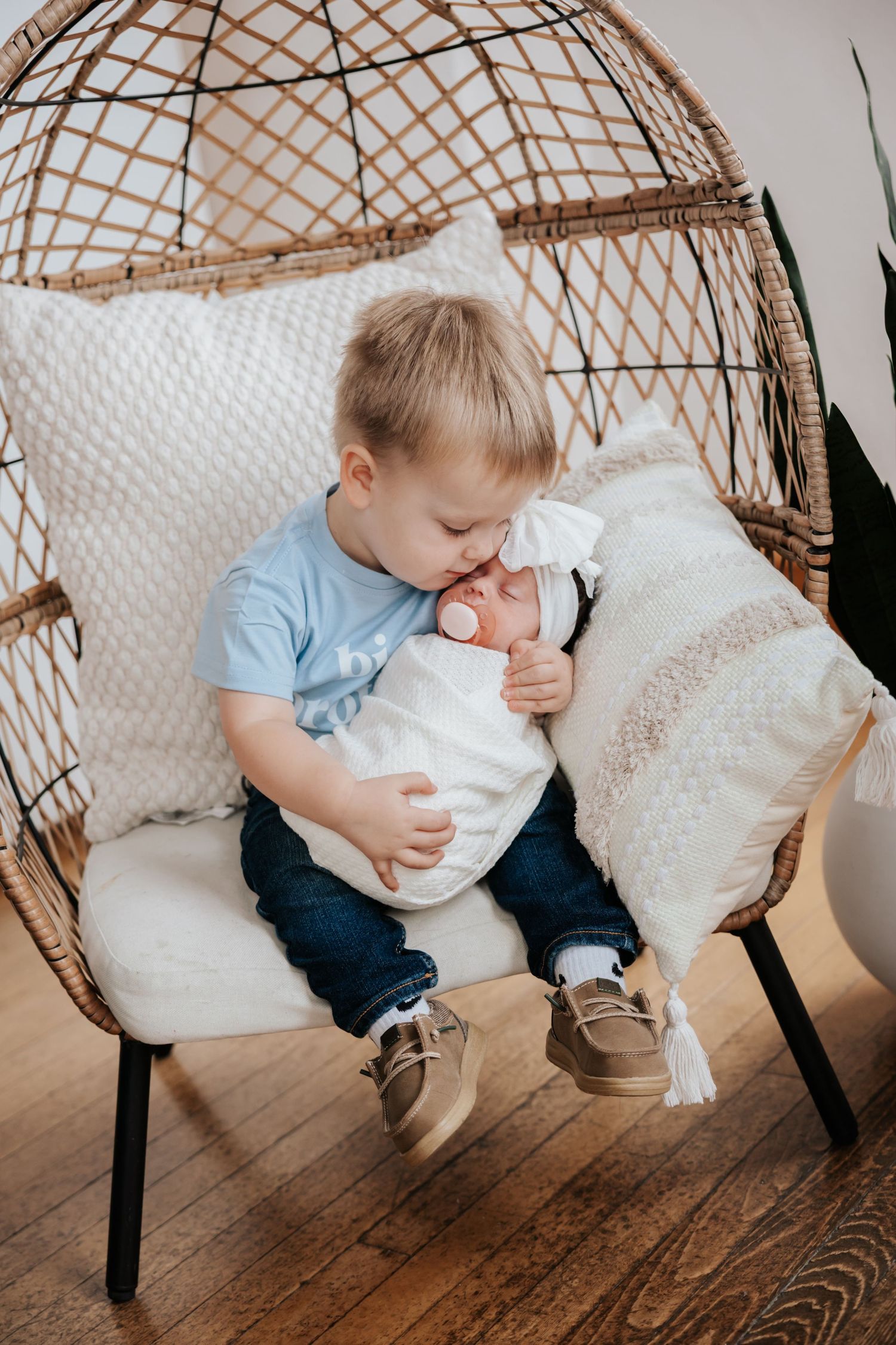 Toddler boy in a blue shirt gently kisses his swaddled newborn sister's forehead while sitting in a cozy wicker egg chair.