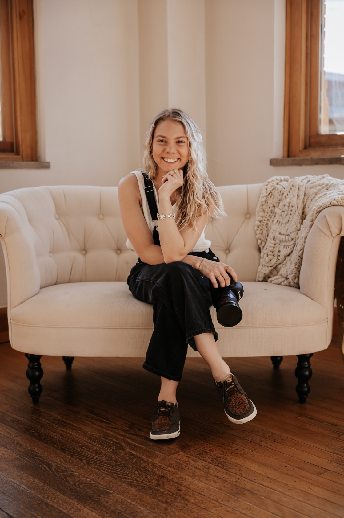 Smiling woman with long blonde hair sits on a cream tufted sofa holding a camera in a bright studio.