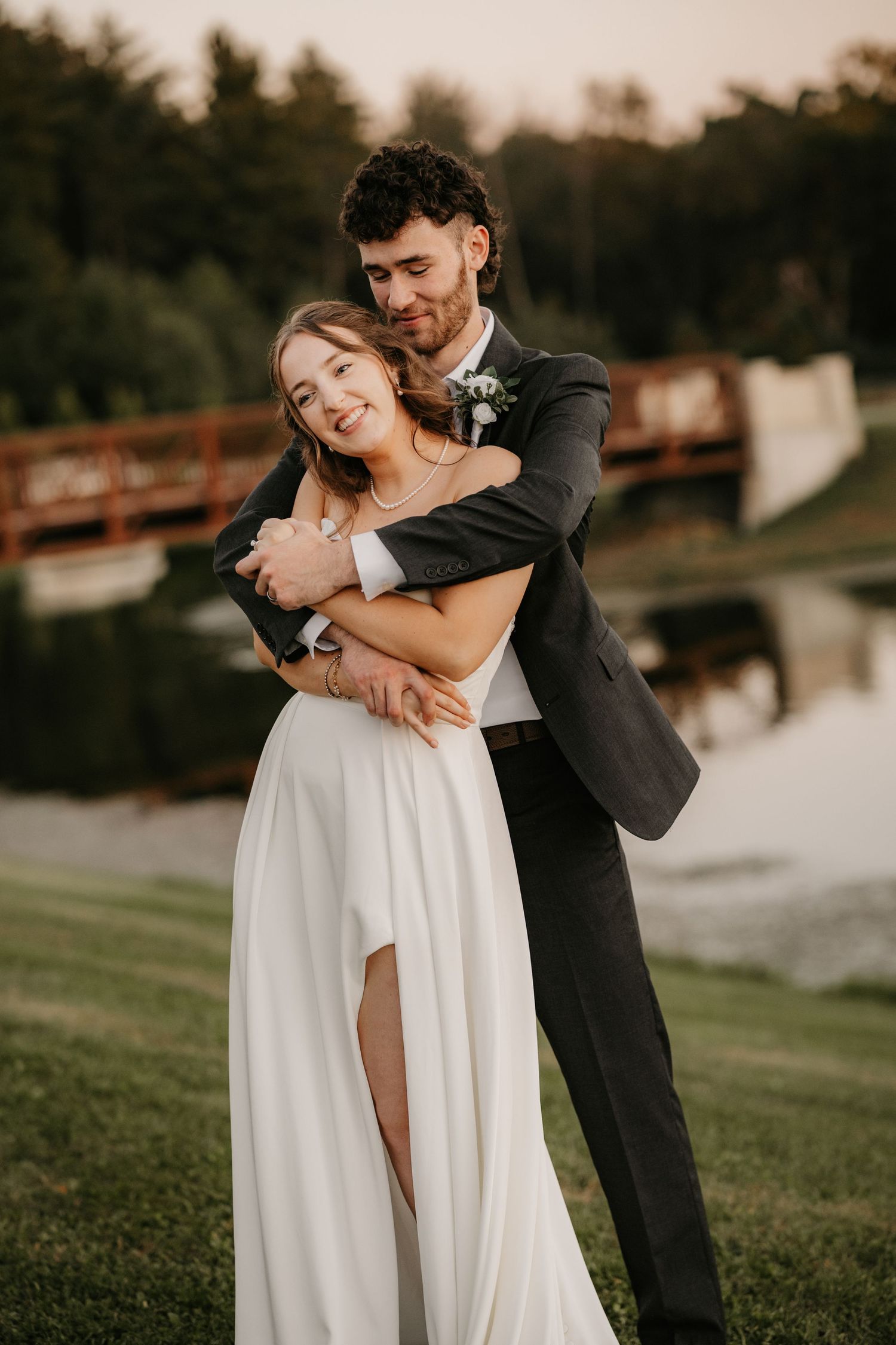 Joyful bride in a white gown with a leg slit is wrapped in her groom's arms on the lawn beside a pond and wooden bridge.