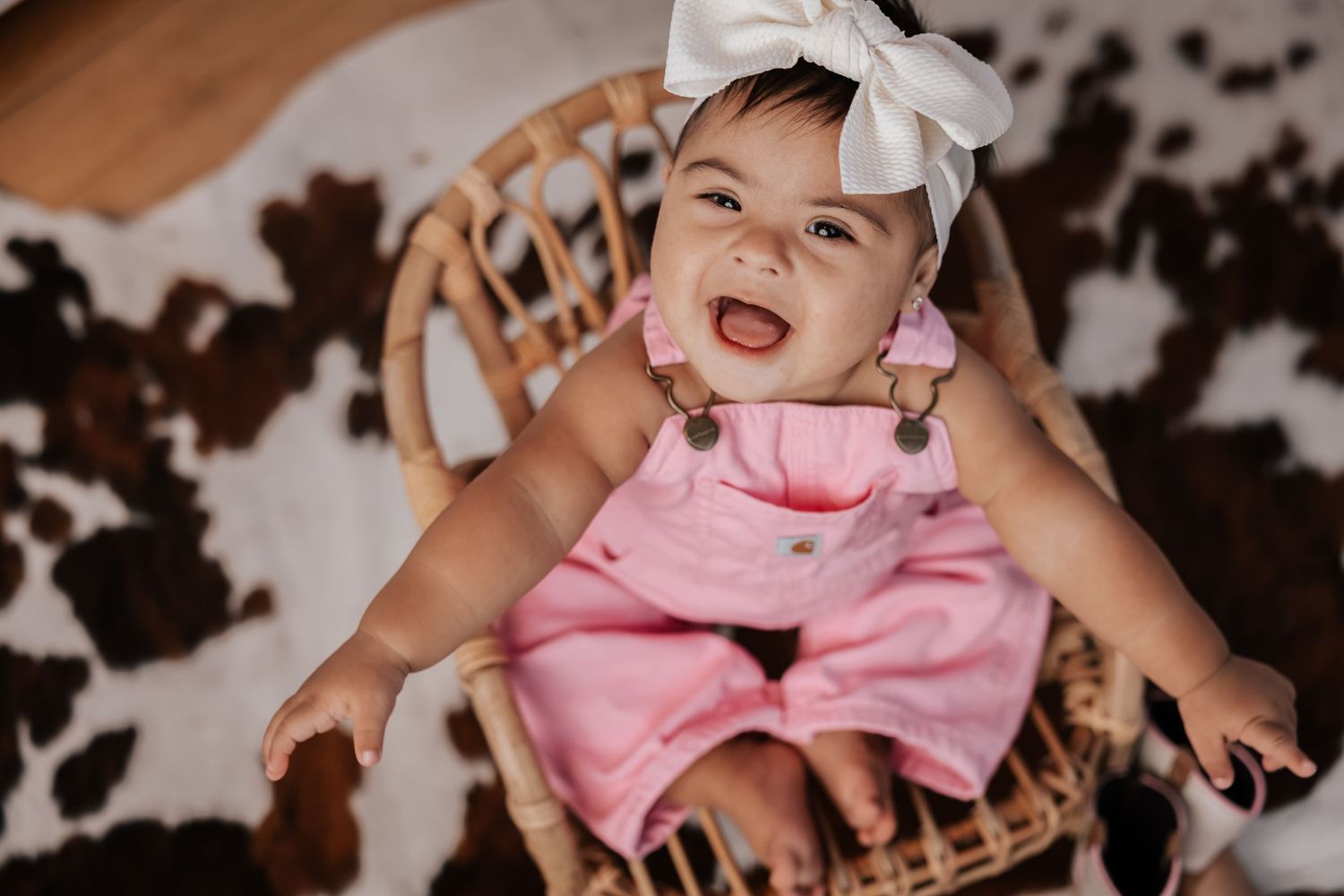 Overhead view of a smiling baby in pink overalls and a white bow sitting in a rattan chair on a cowhide rug.