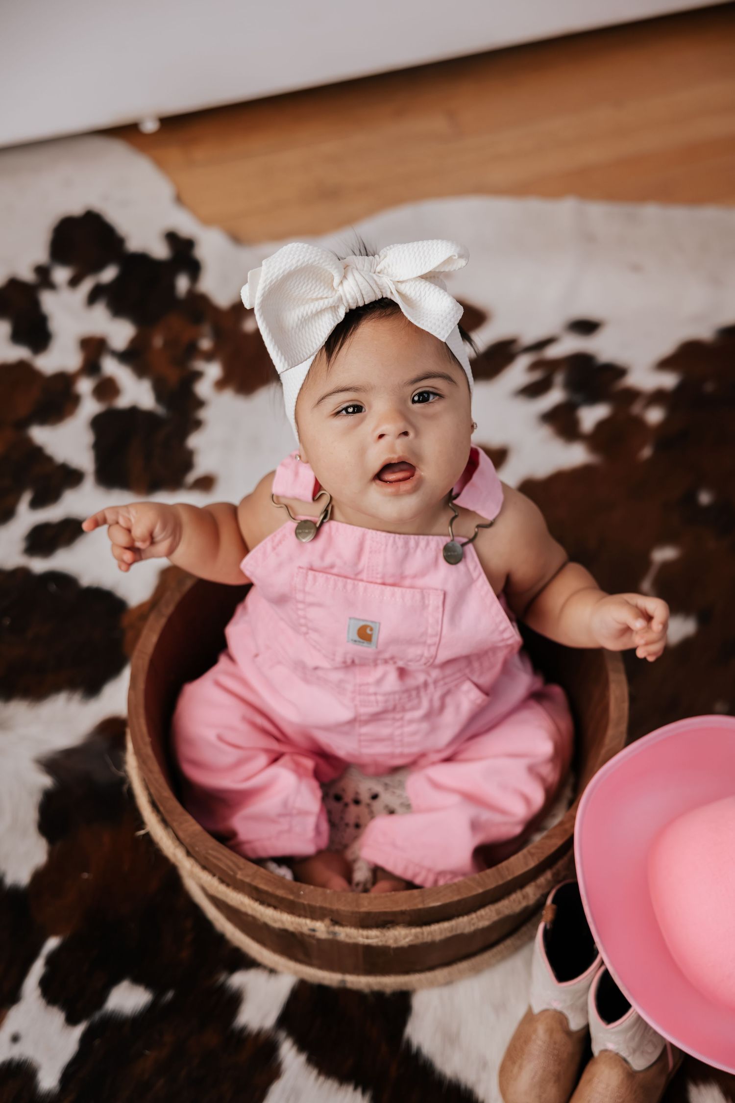 Baby girl in pink overalls and a big white bow looks up from a wooden barrel on a cowhide rug next to cowgirl boots.