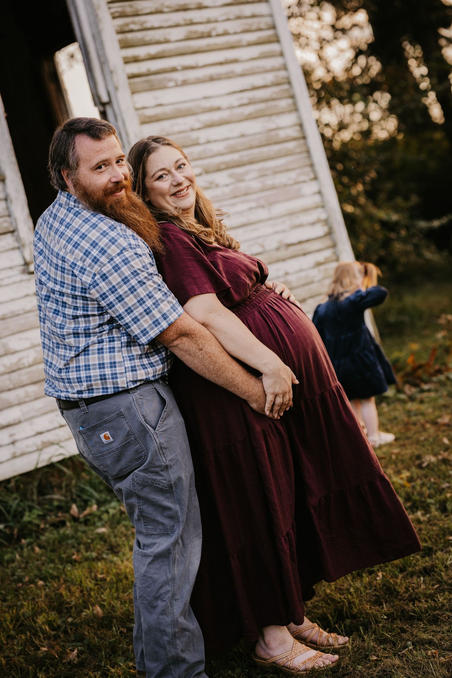 Smiling expecting couple embraces beside a weathered white barn while their toddler daughter plays in the grass behind them.