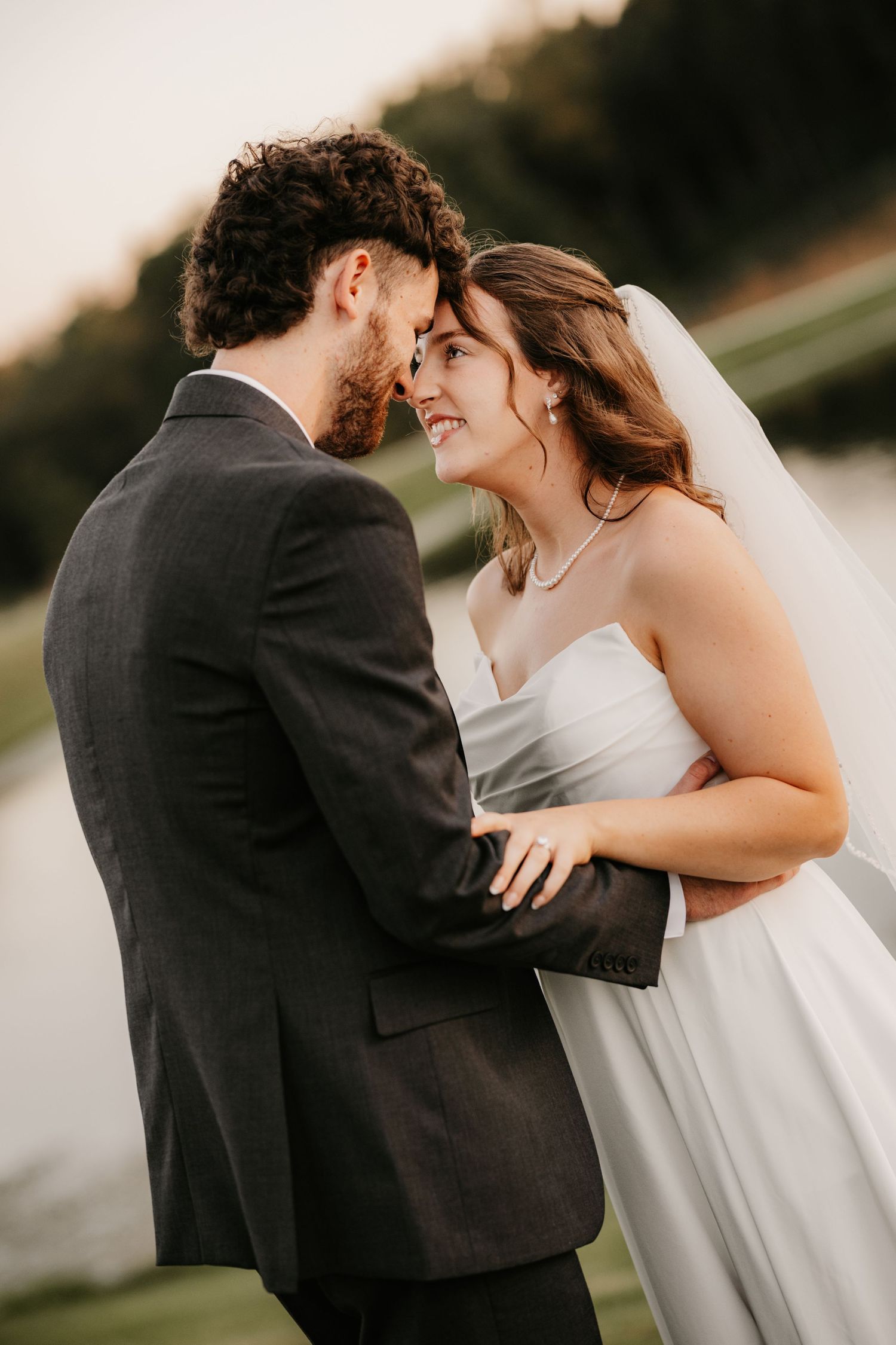 Smiling bride and groom touch foreheads in an affectionate embrace beside a calm lake at sunset.