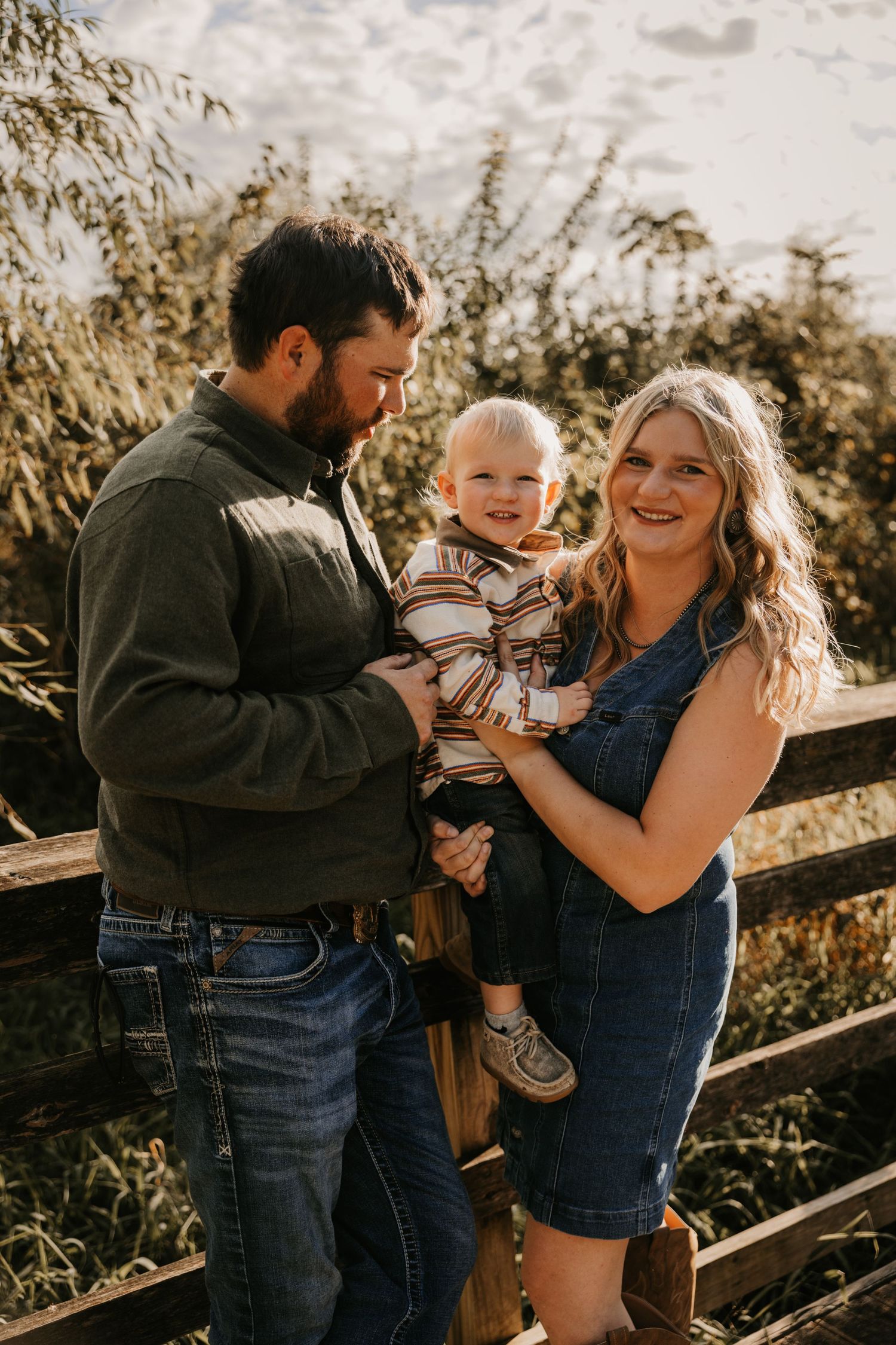 A bearded father and smiling mother in a denim dress hold their toddler son beside a wooden fence in a warm outdoor family portrait.