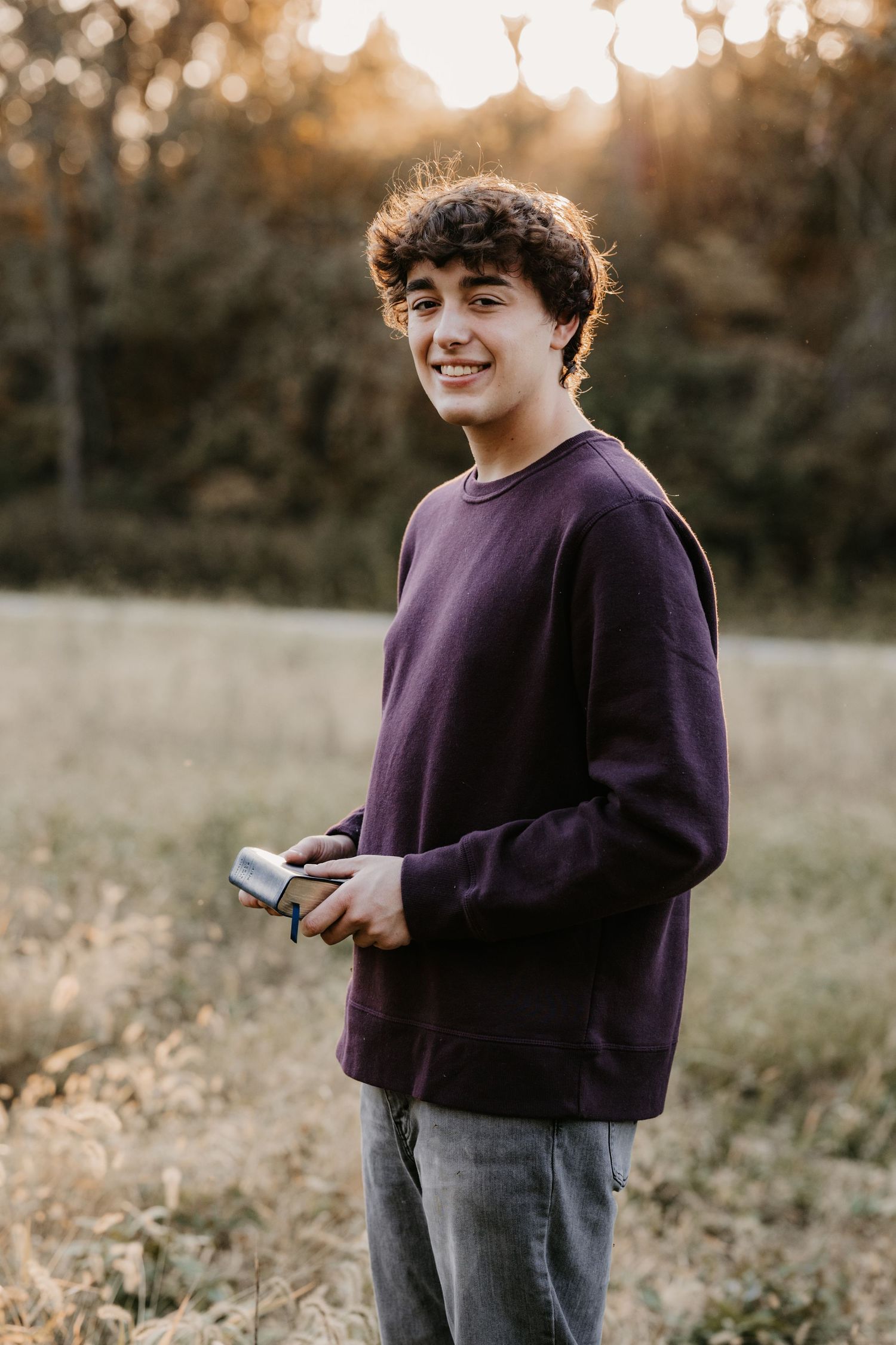 Smiling young man in a maroon sweatshirt holds a small camera in a grassy field as golden sunset light glows behind him.