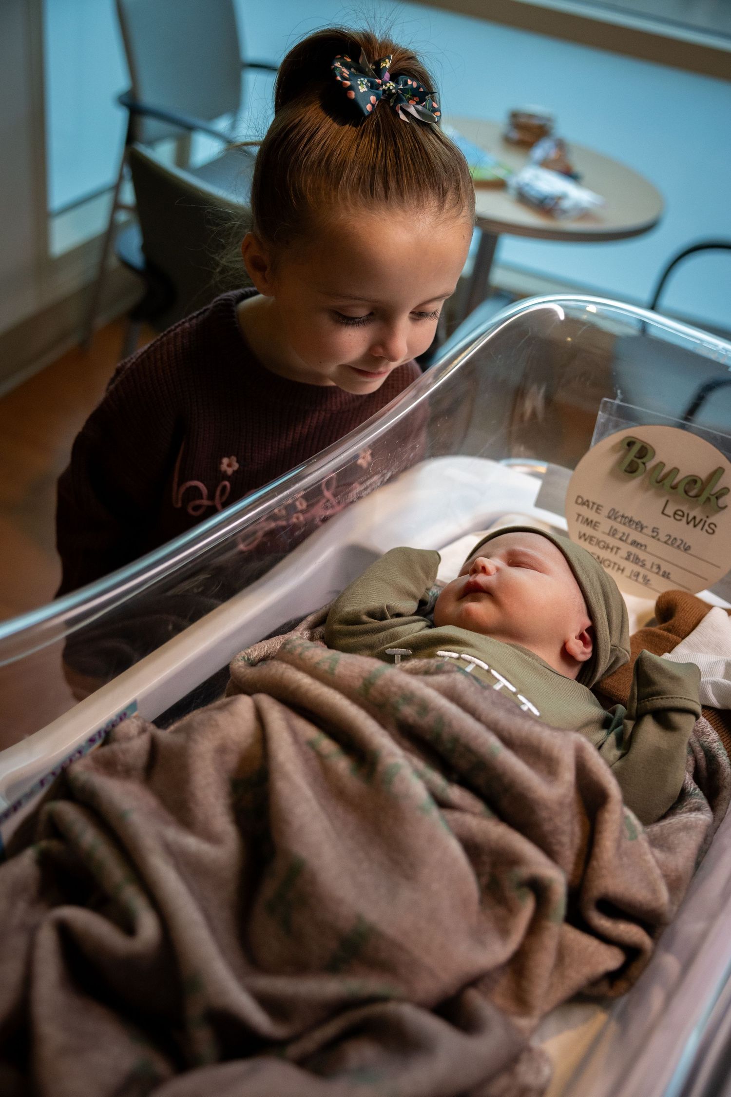 A young girl with a bow in her hair gazes lovingly at her sleeping newborn sibling in a hospital bassinet.