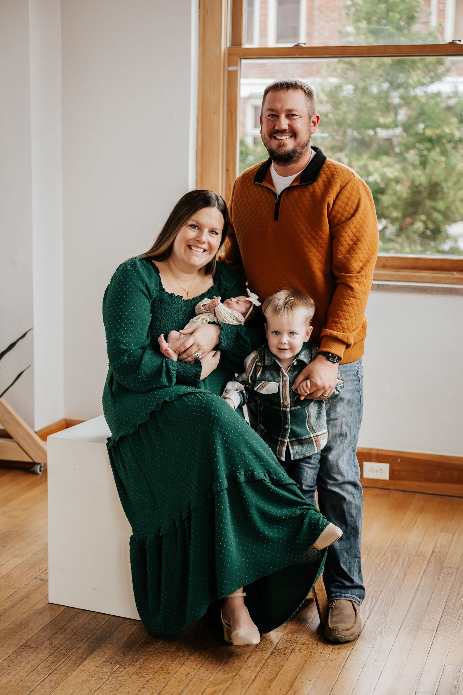 Family portrait of parents and their toddler son posing with a newborn baby girl, mom in green dress and dad in rust sweater.