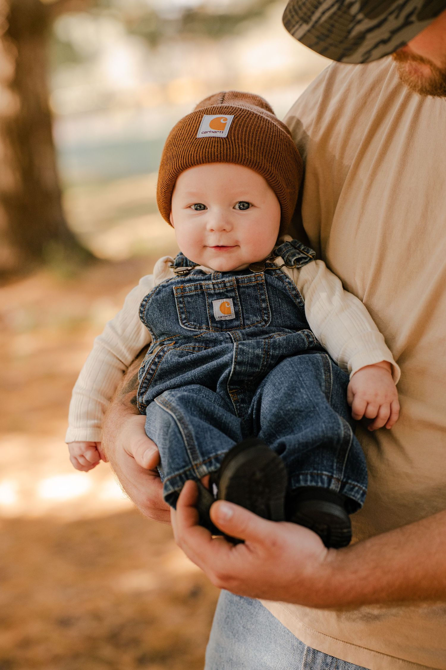 Smiling baby in a brown Carhartt beanie and denim overalls is held in a dad's arms during an outdoor autumn family photo session.