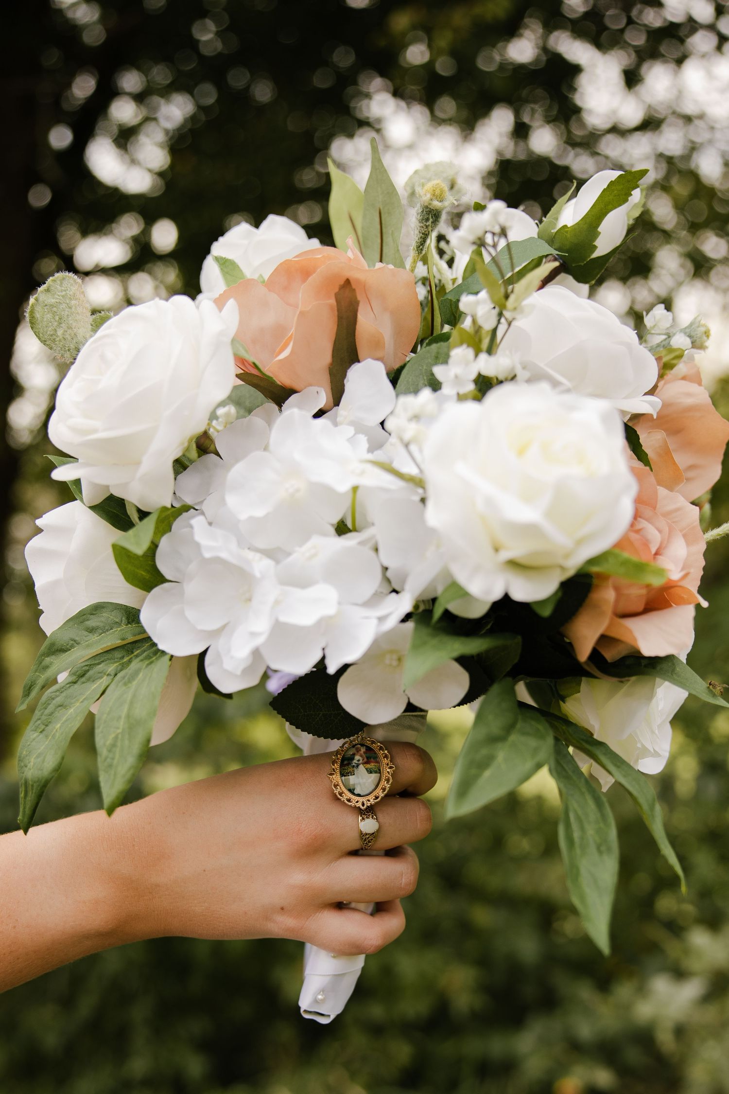 Hand holding a bridal bouquet of white and peach roses with greenery and a vintage cameo ring.