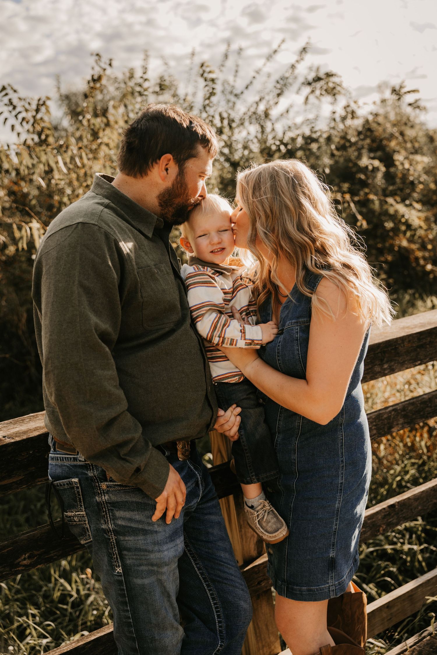 Parents tenderly kiss their toddler's cheeks beside a wooden fence in a heartfelt outdoor family portrait with warm autumn tones.