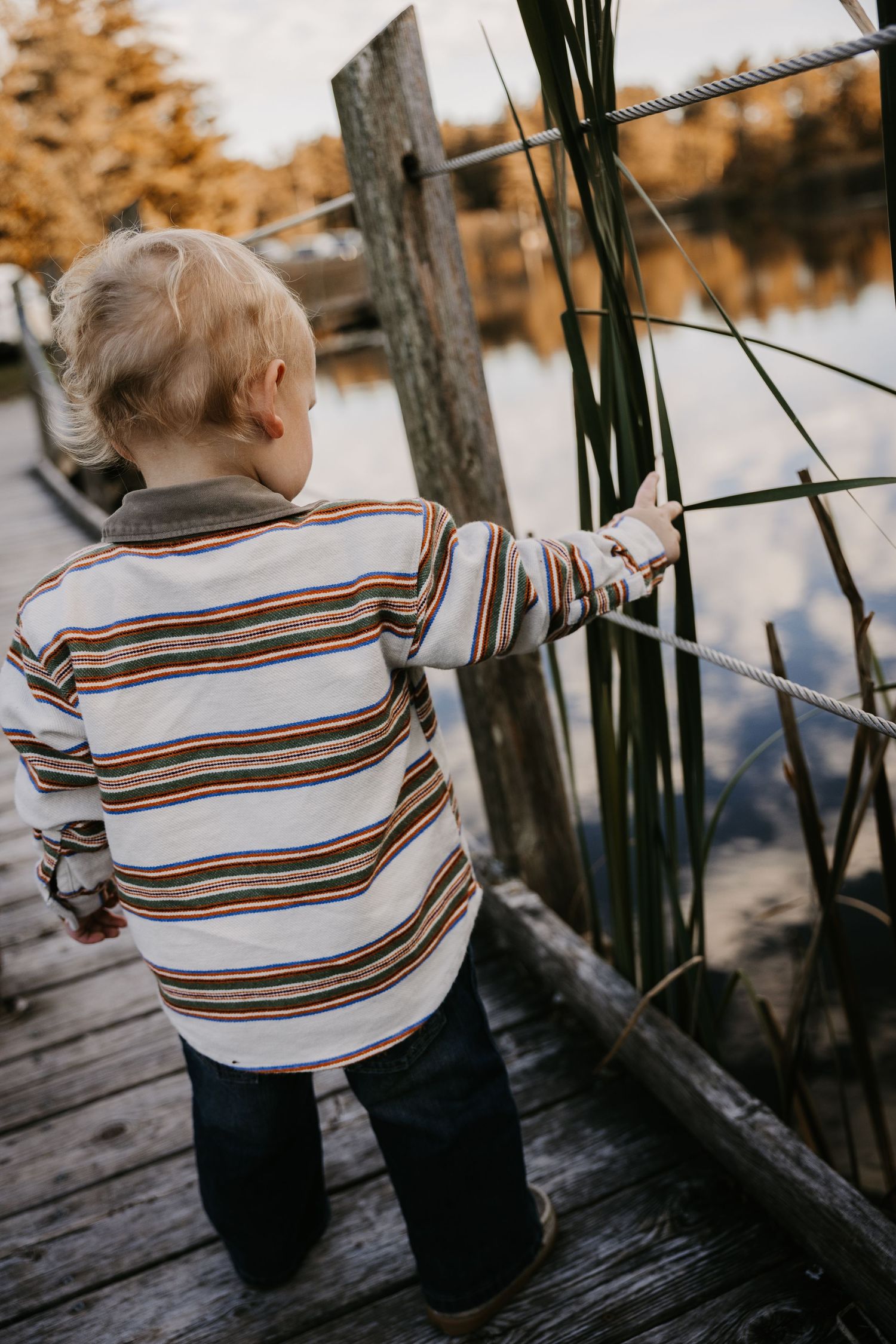 A toddler in a striped sweater reaches for a rope railing on a wooden dock overlooking a calm lake and golden autumn trees.