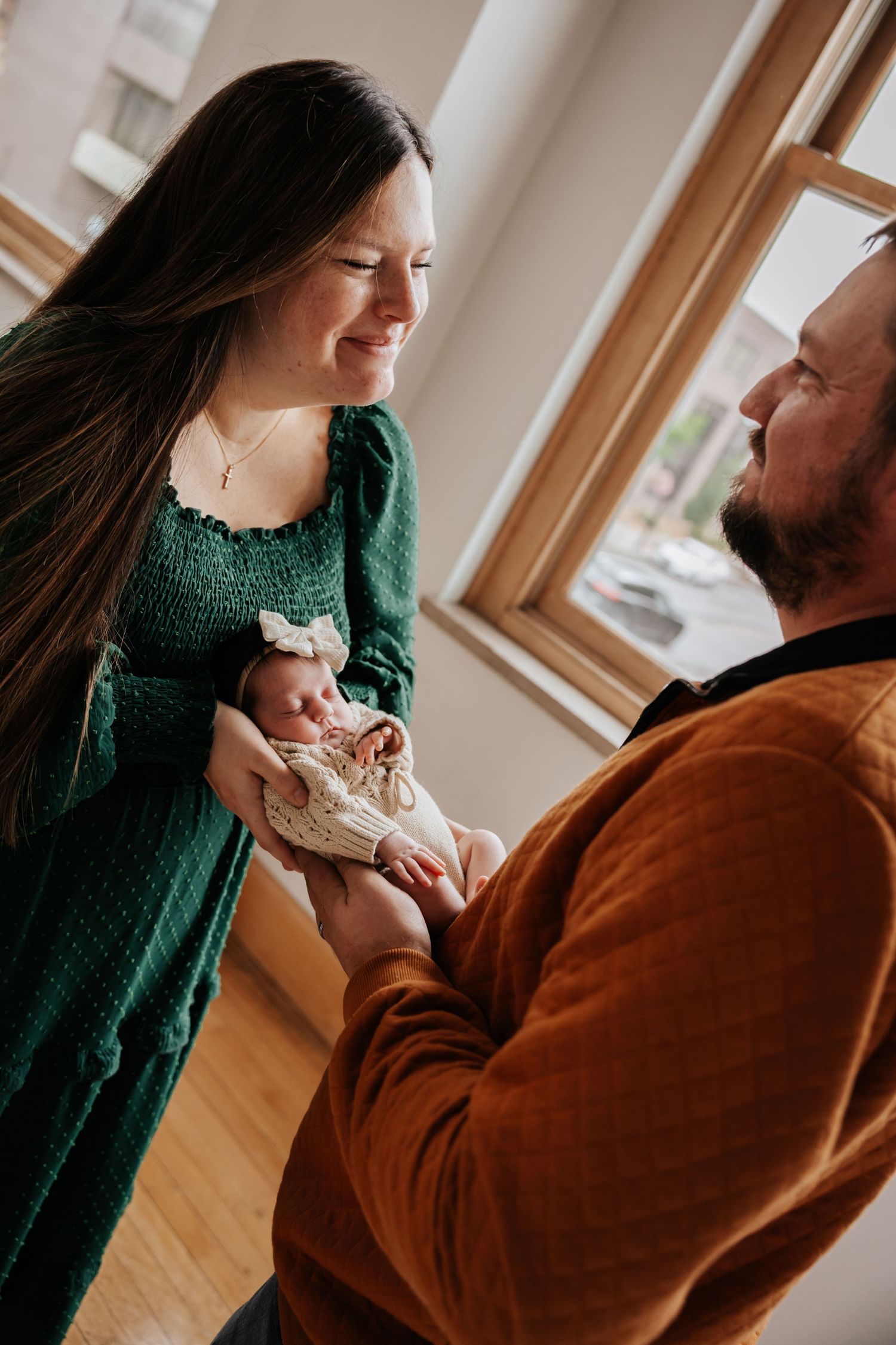 Smiling mom holds her bow-wearing newborn while dad in a rust quilted pullover watches lovingly near a bright window.