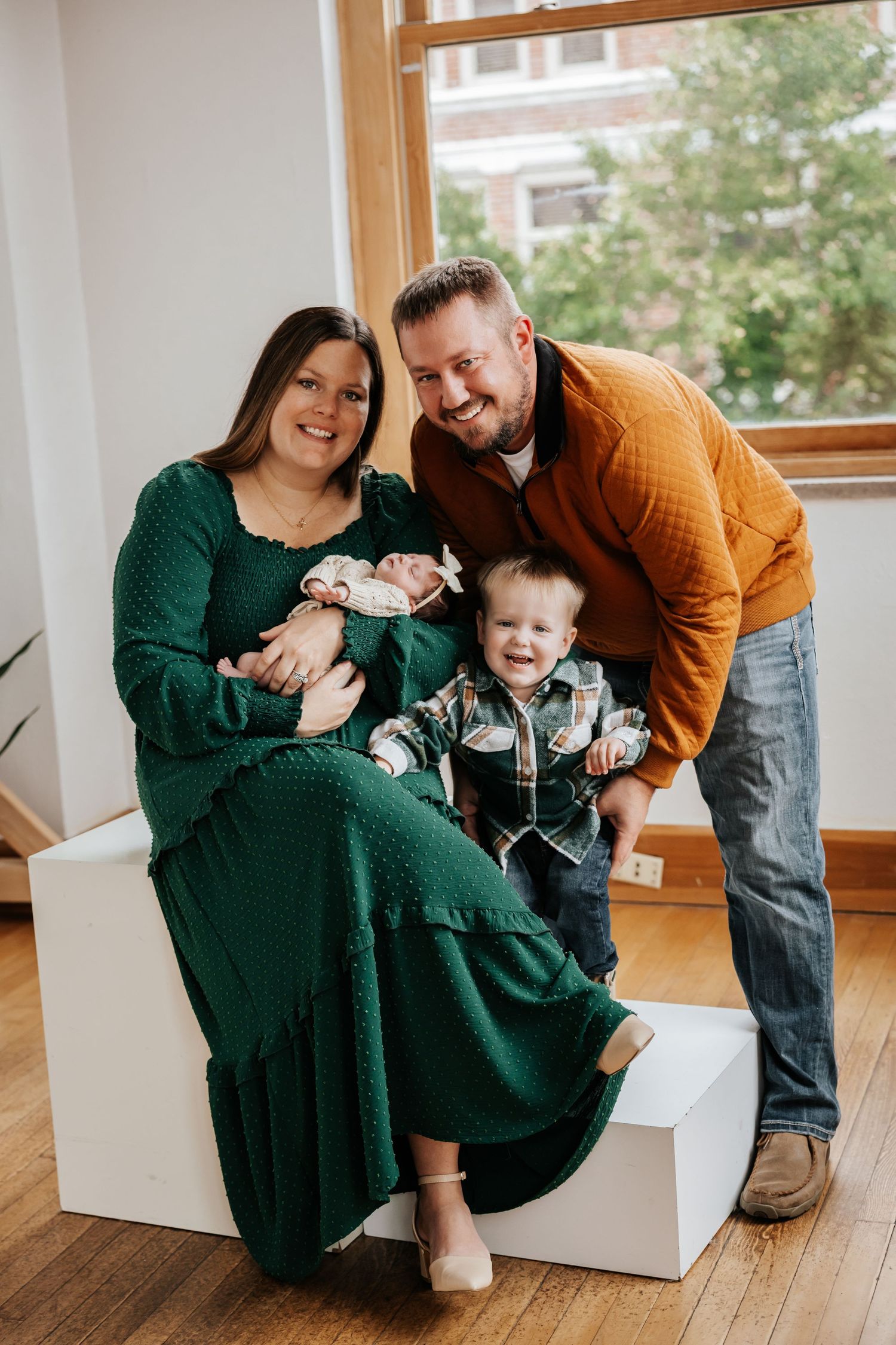 A smiling family of four poses by a sunny window, with parents in green and orange holding a toddler and newborn baby.
