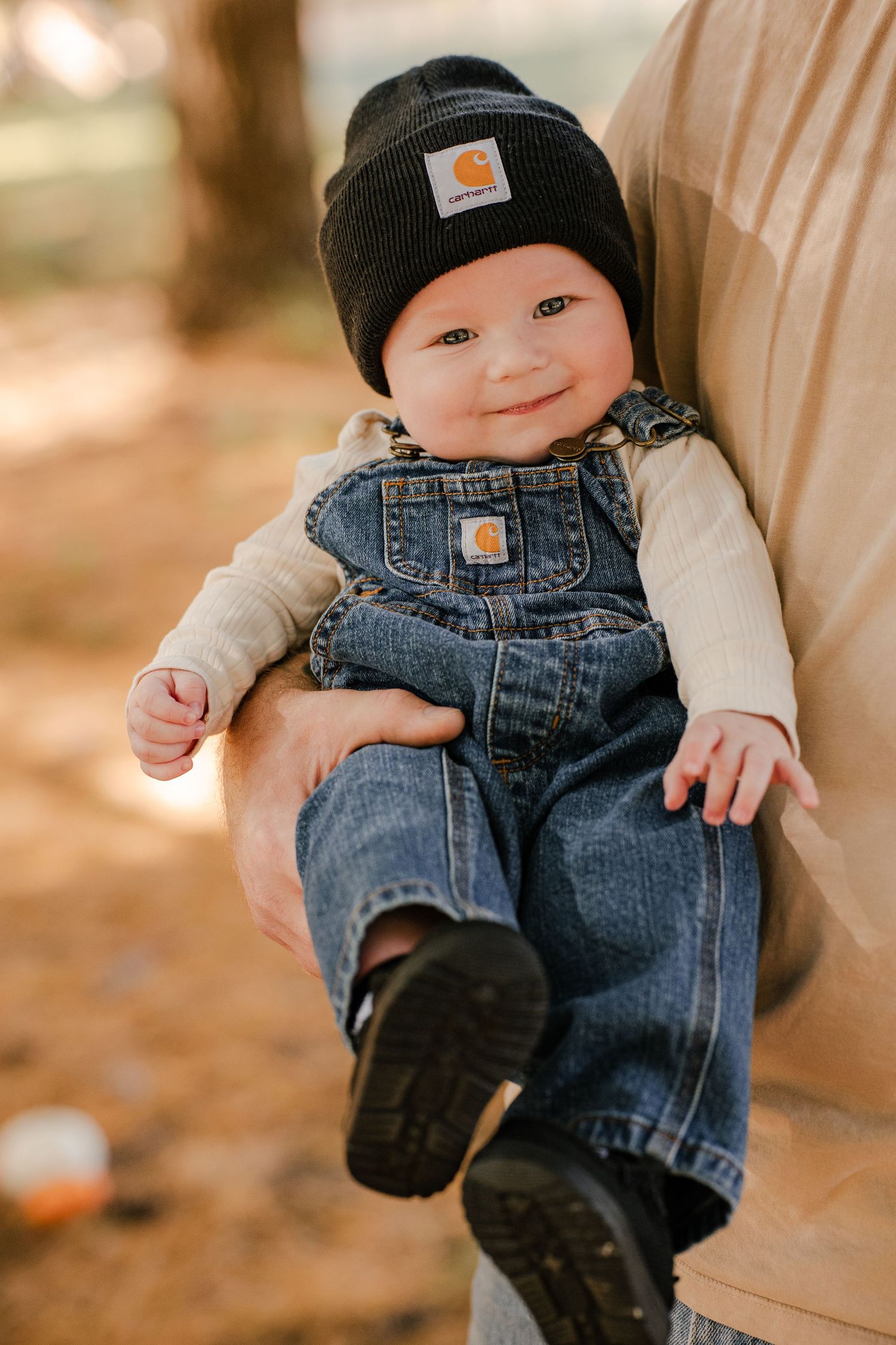 Smiling baby in a black Carhartt beanie and denim overalls is held sideways by a parent in a warm, sunlit autumn forest.