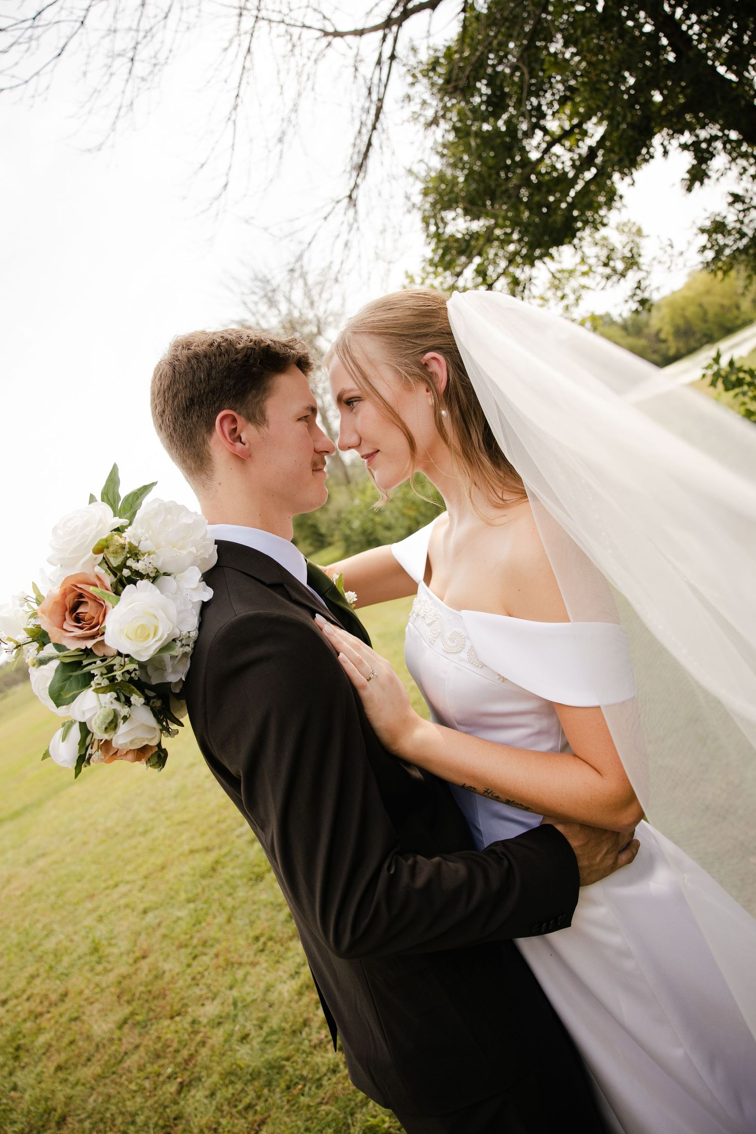 Bride and groom share a tender moment nose to nose outdoors with a floral bouquet and flowing veil.