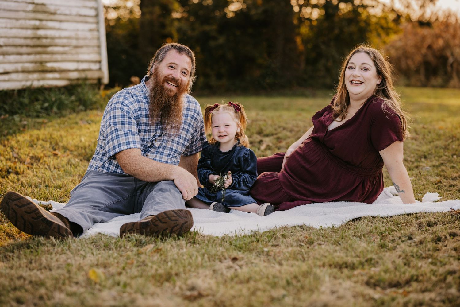 Bearded dad, pregnant mom and their toddler daughter sit together on a blanket in the grass for a warm outdoor maternity family photo.