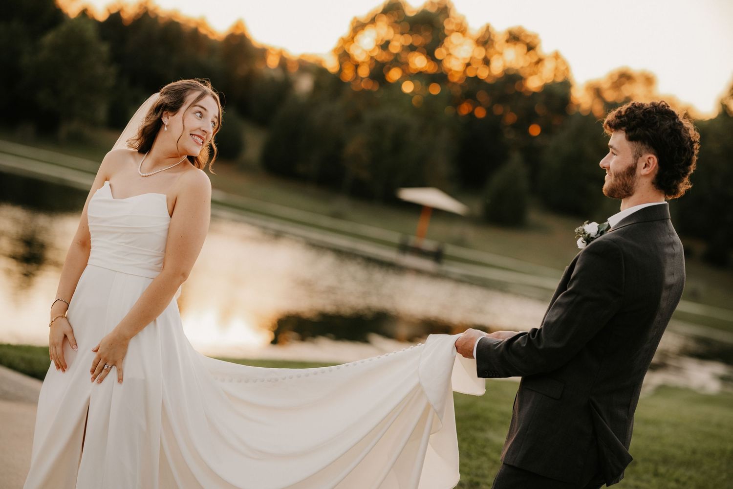 Smiling bride holds her flowing white wedding gown while the groom looks on beside a pond at sunset.