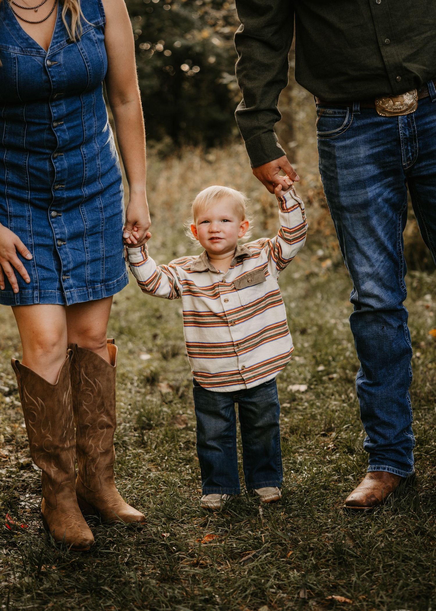 A toddler in a striped shirt holds hands with his parents while walking through a grassy field, with cowboy boots and jeans visible.