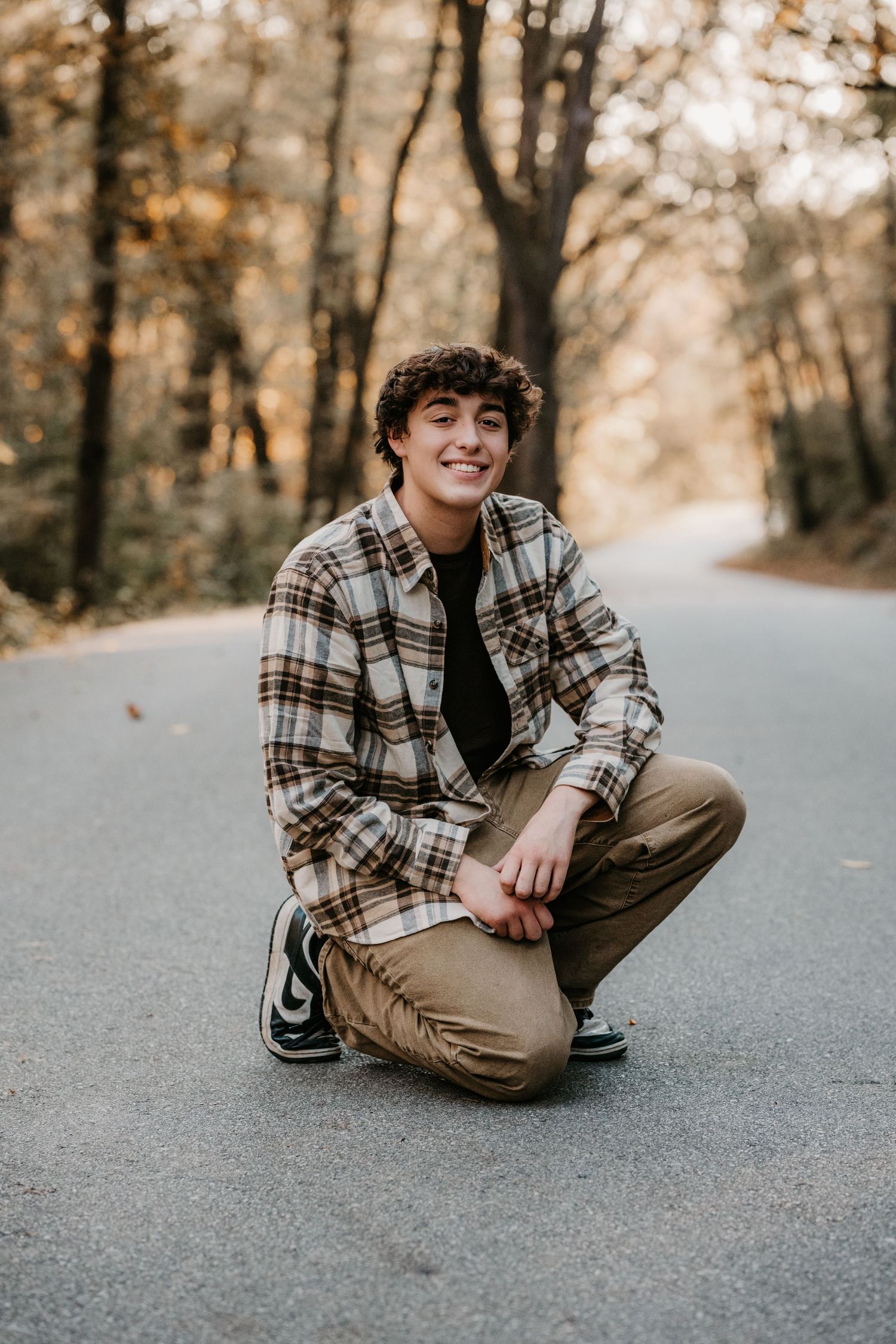 Smiling young man with curly hair crouches on a quiet tree-lined road in a plaid flannel shirt, khaki pants, and sneakers.