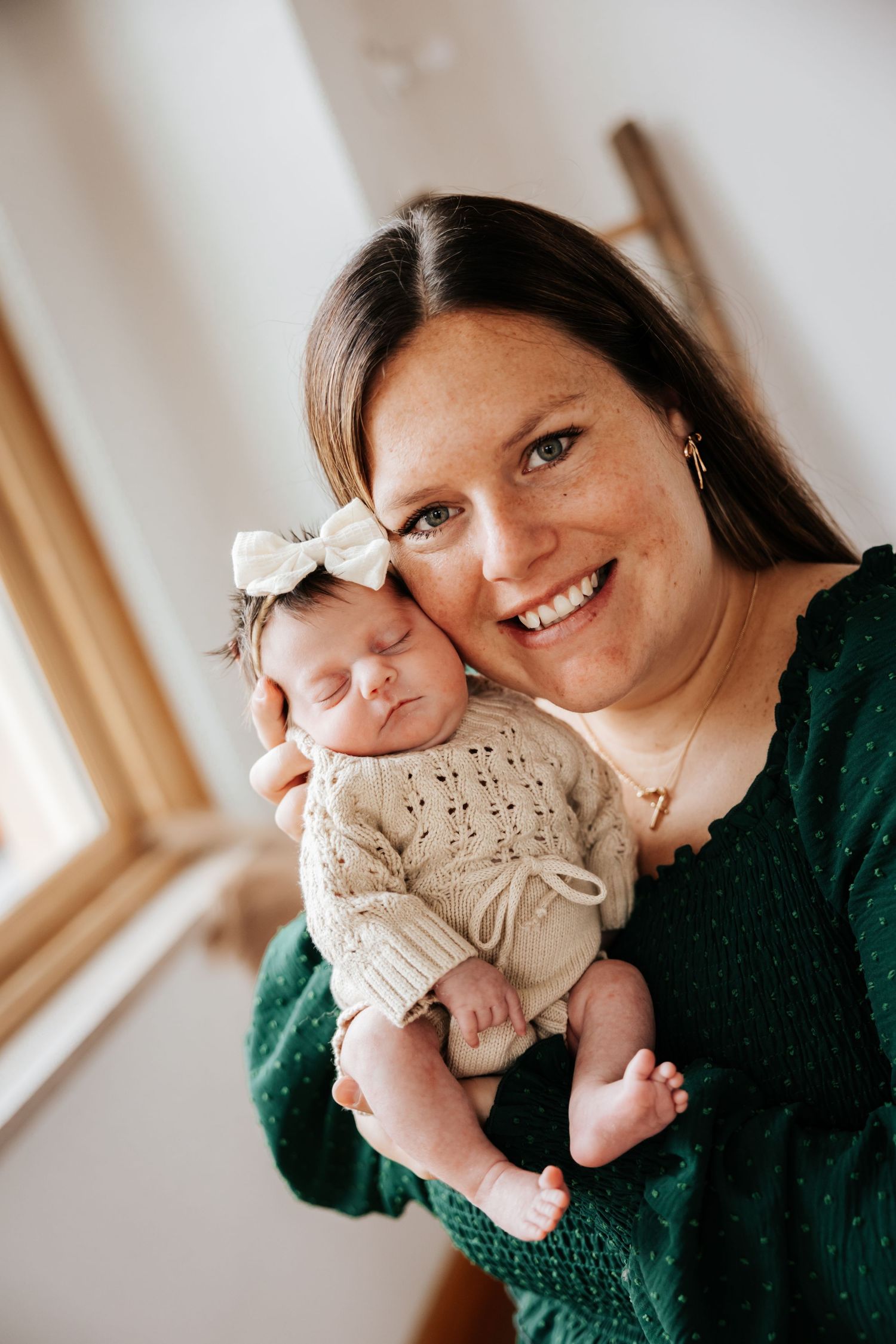 Smiling mother in a green polka dot dress snuggles her sleeping newborn daughter wearing a cream bow headband.