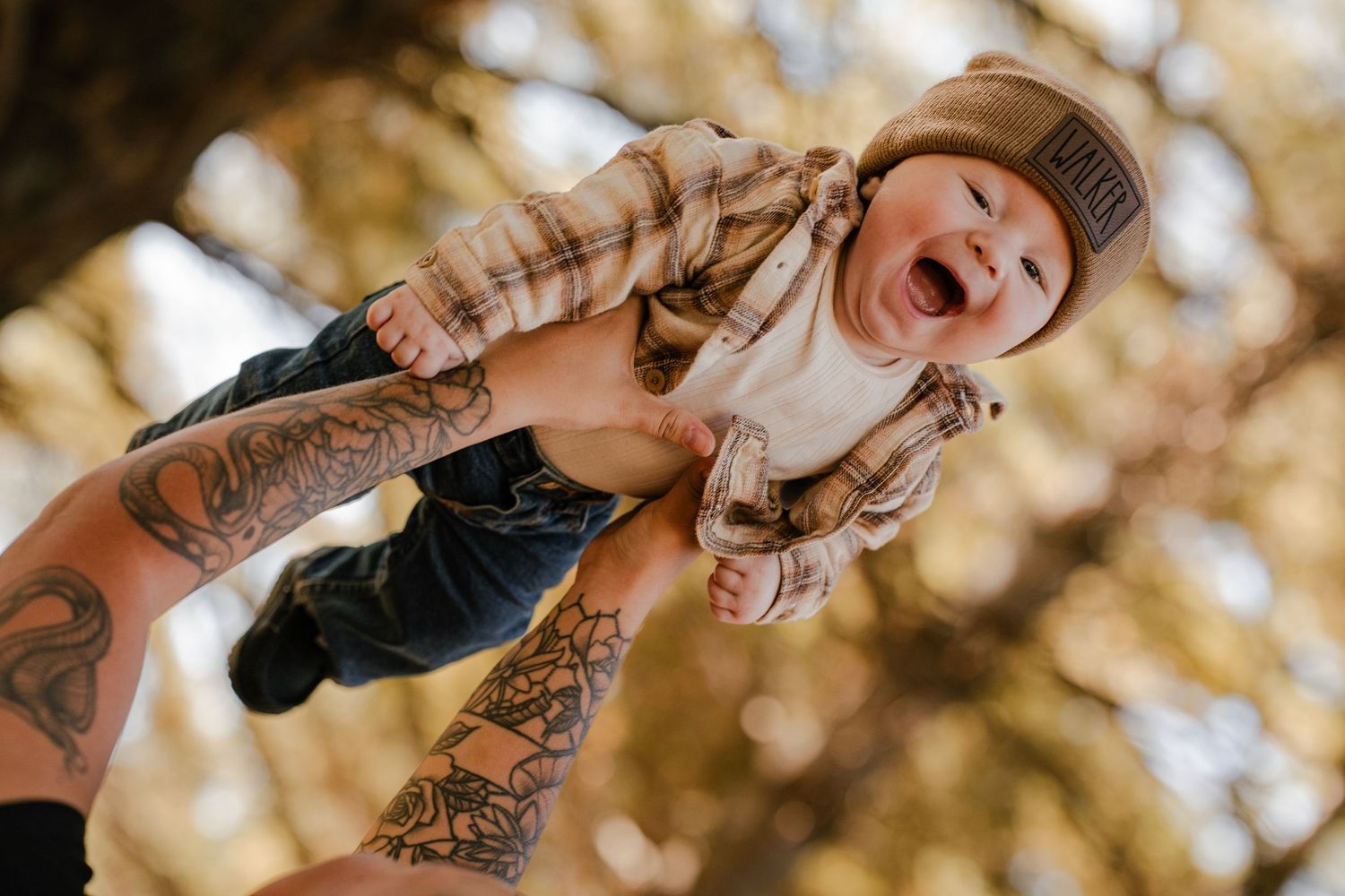 Joyful baby in a plaid flannel shirt and beanie is lifted high into the air by tattooed arms against golden autumn trees.