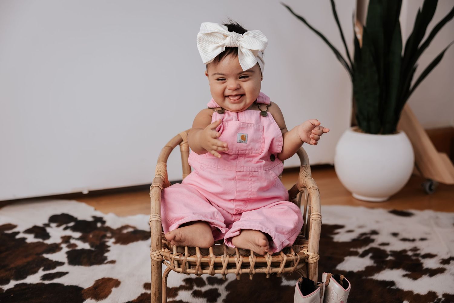 Happy baby in pink overalls and a cream bow headband sits in a small rattan chair next to a potted plant and cowboy boots.