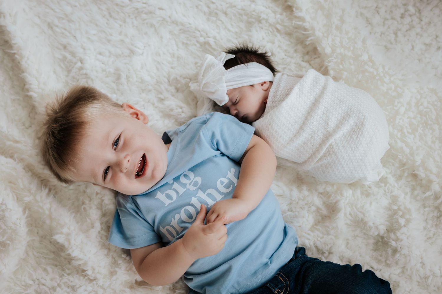 Laughing toddler in a big brother shirt lies on a white fur blanket next to his swaddled newborn sister wearing a headband.
