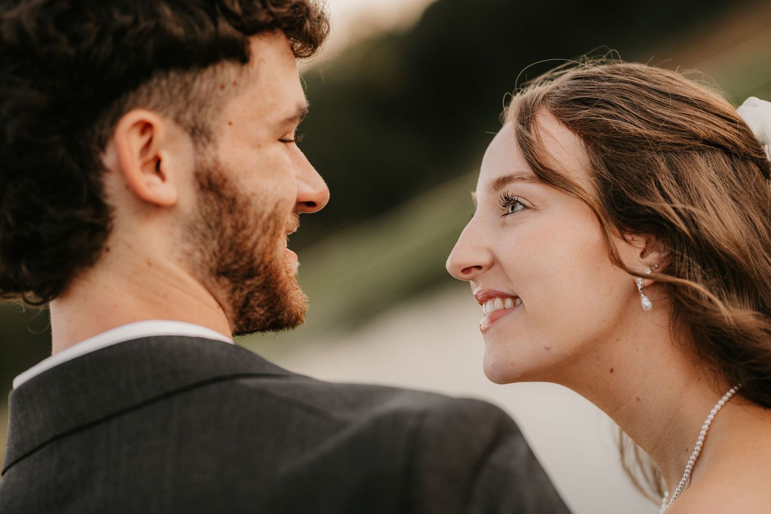 Close-up of a bride and groom smiling at each other, framed by soft evening light and a blurred outdoor background.