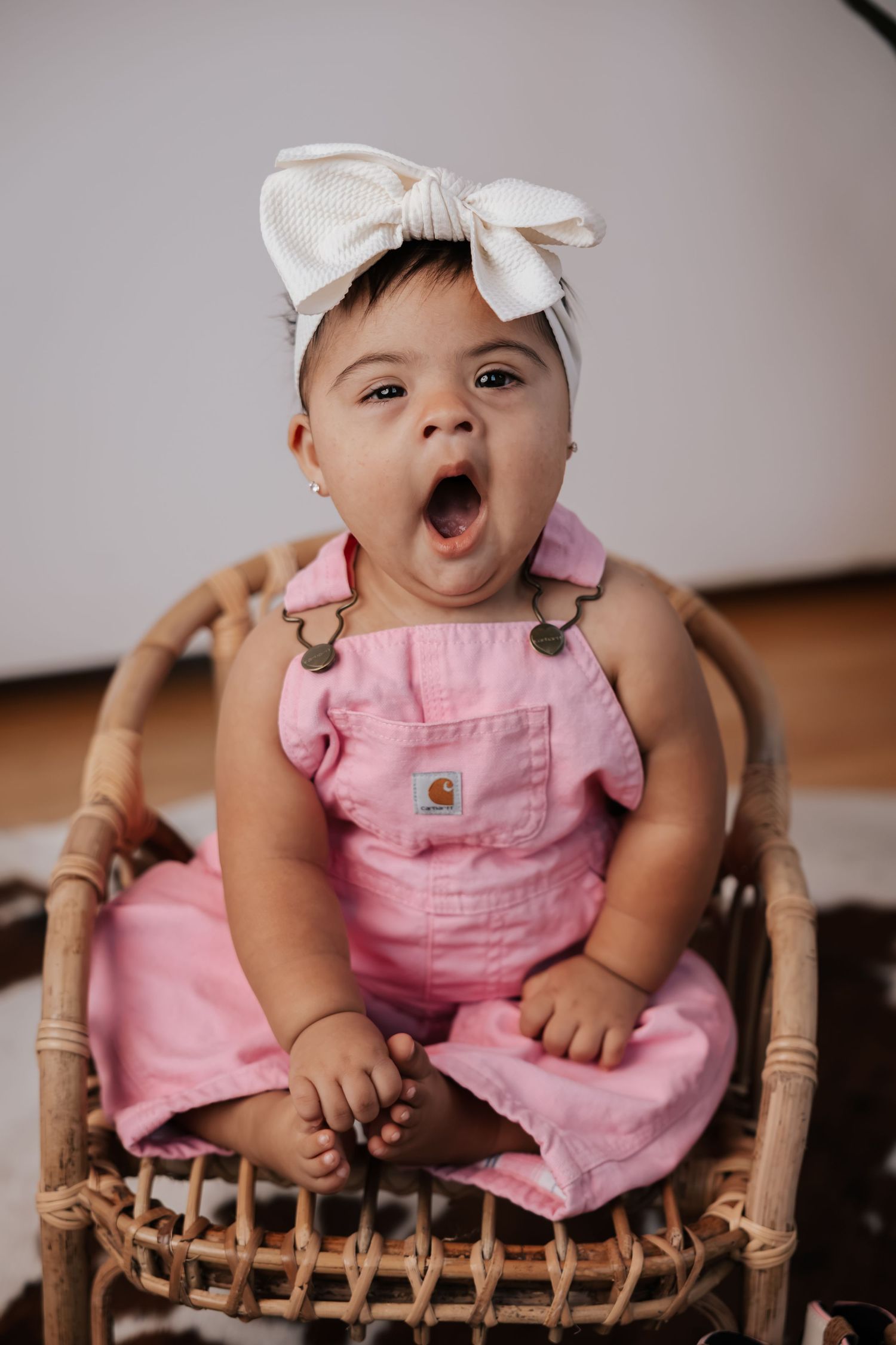 Baby in pink overalls and a cream bow headband yawns widely while seated in a woven rattan chair during a photo session.