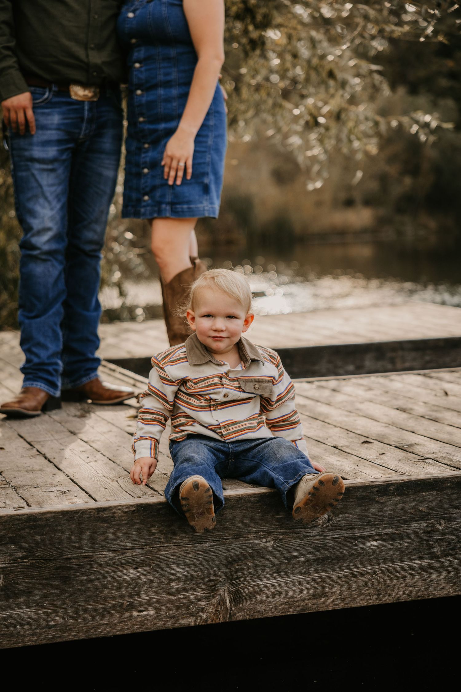 Toddler in a striped shirt and jeans sits on a rustic wooden dock while his parents stand behind him by the water.