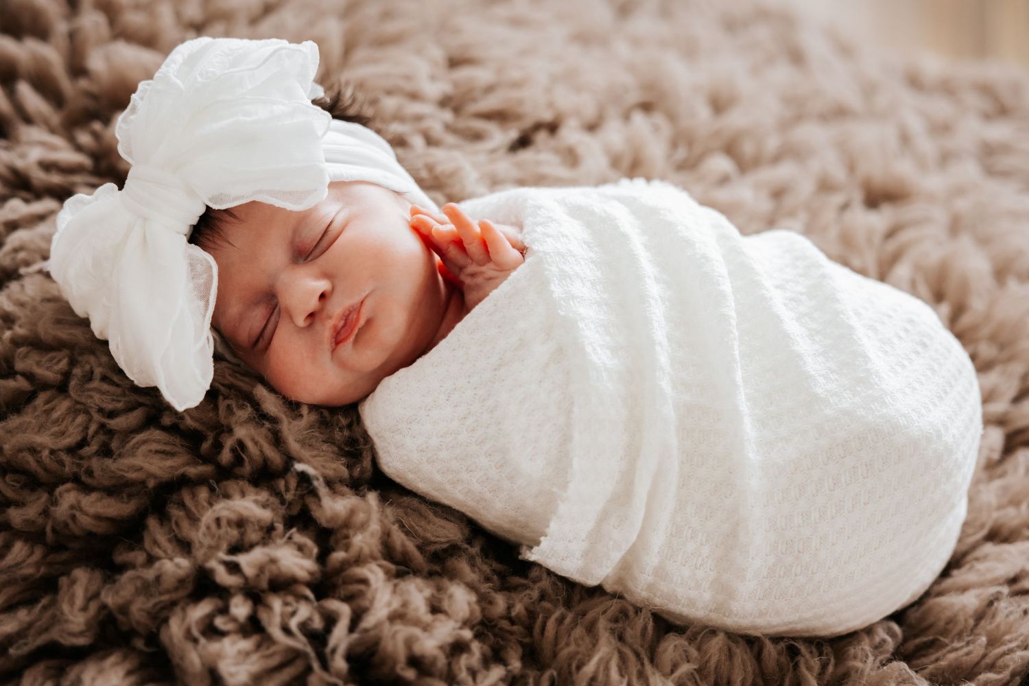 Peaceful newborn girl in a white swaddle and oversized white bow lies on a textured brown fur rug with her tiny hand showing.