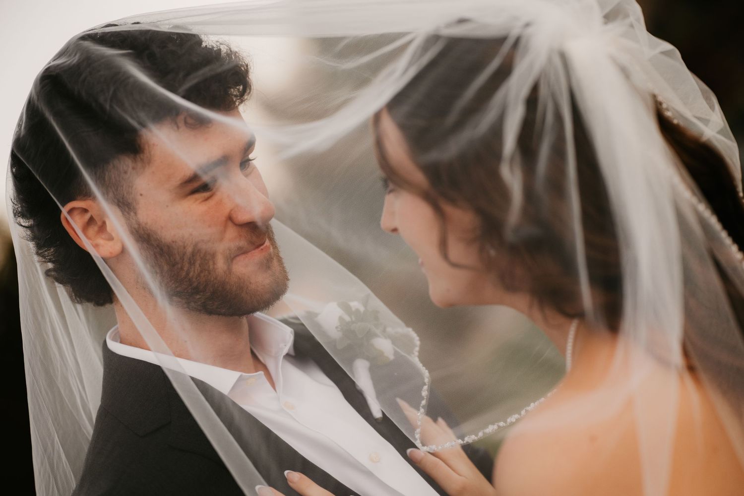 Groom smiles at his bride as they share an intimate moment beneath her sheer veil in soft golden light.