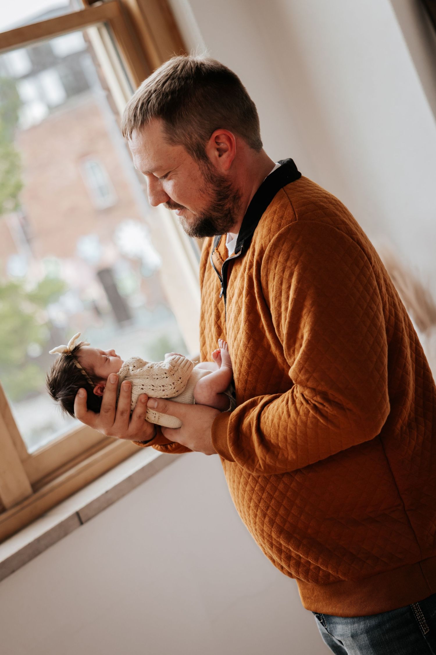 Bearded father in a rust quilted sweater gazes lovingly at his newborn baby cradled in his hands by a window.