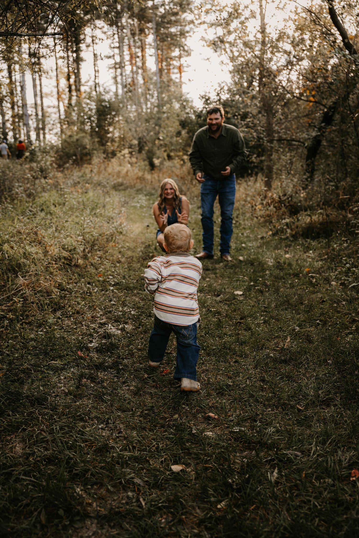 A little boy toddles along a grassy forest trail toward his mother and father, who wait with open arms during a family photo session.