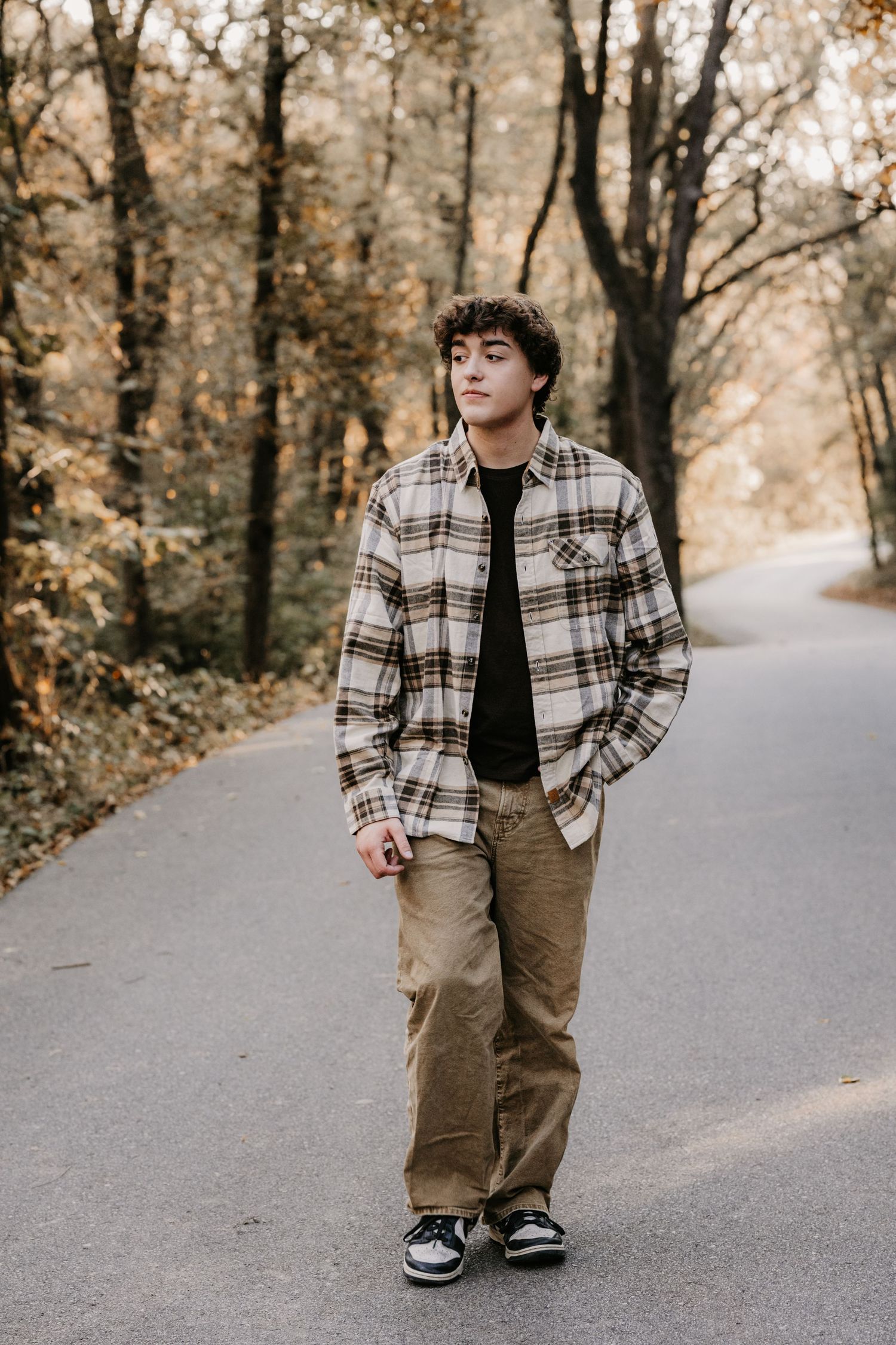 Young man in a plaid flannel overshirt and khaki pants walks down a winding road through autumn woods with hands in pockets.