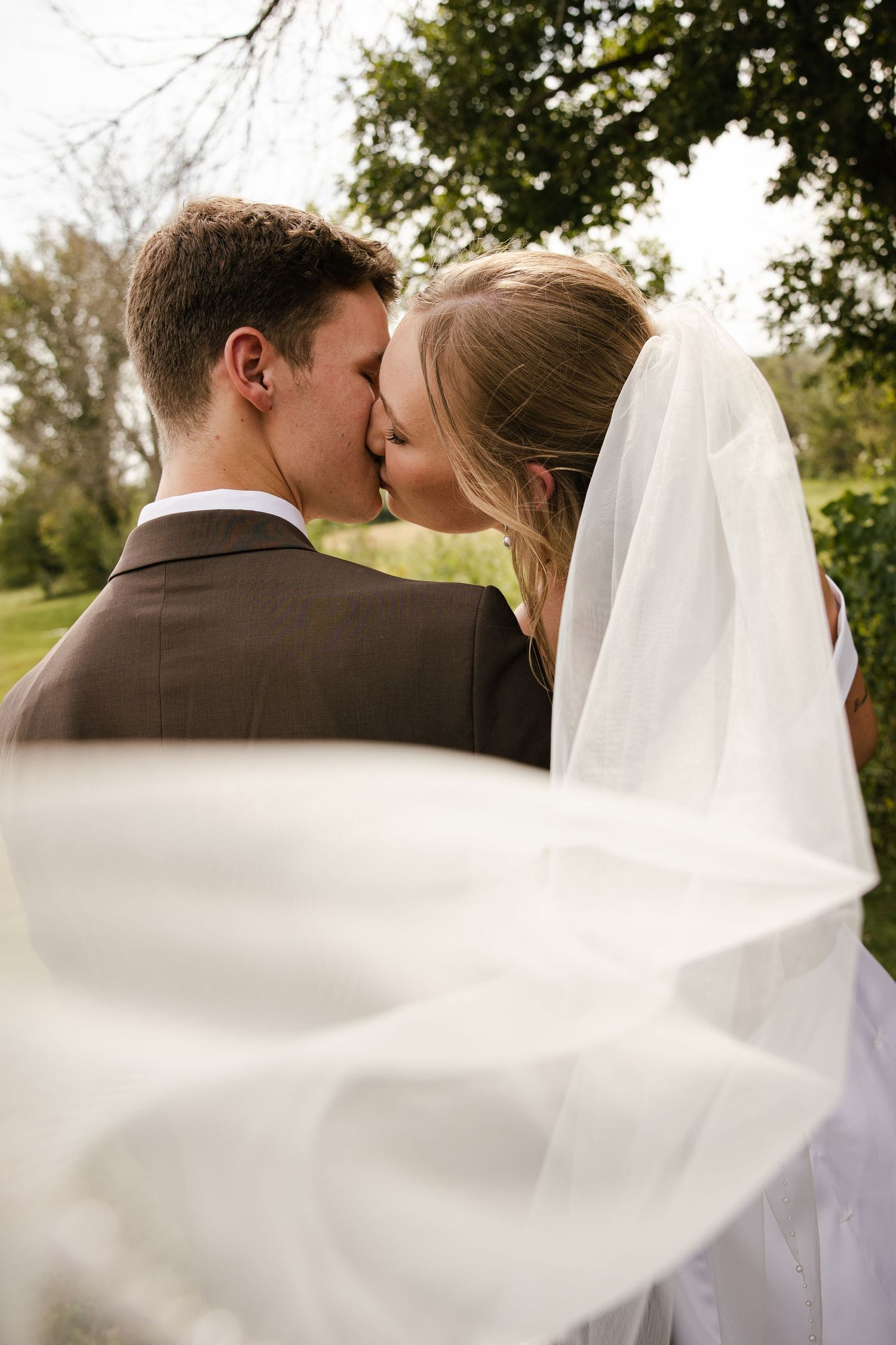 Bride and groom share a romantic kiss outdoors as her sheer wedding veil blows in the breeze.