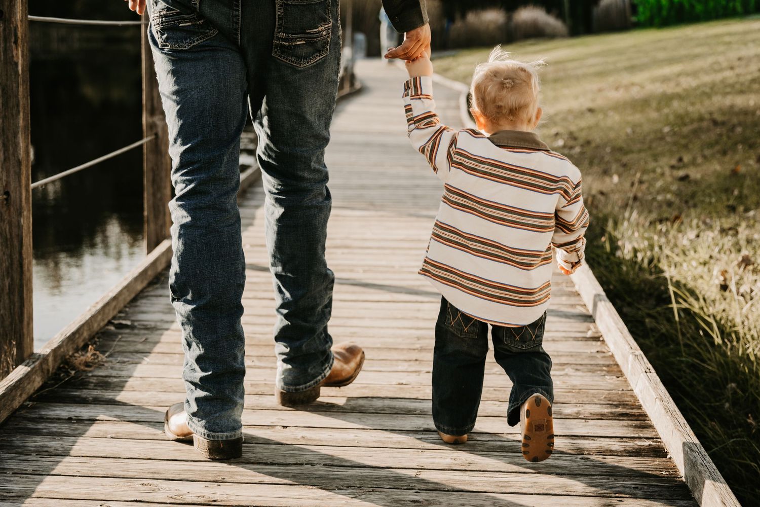 A toddler in a striped sweater walks along a wooden boardwalk beside a lake while holding an adult's hand.