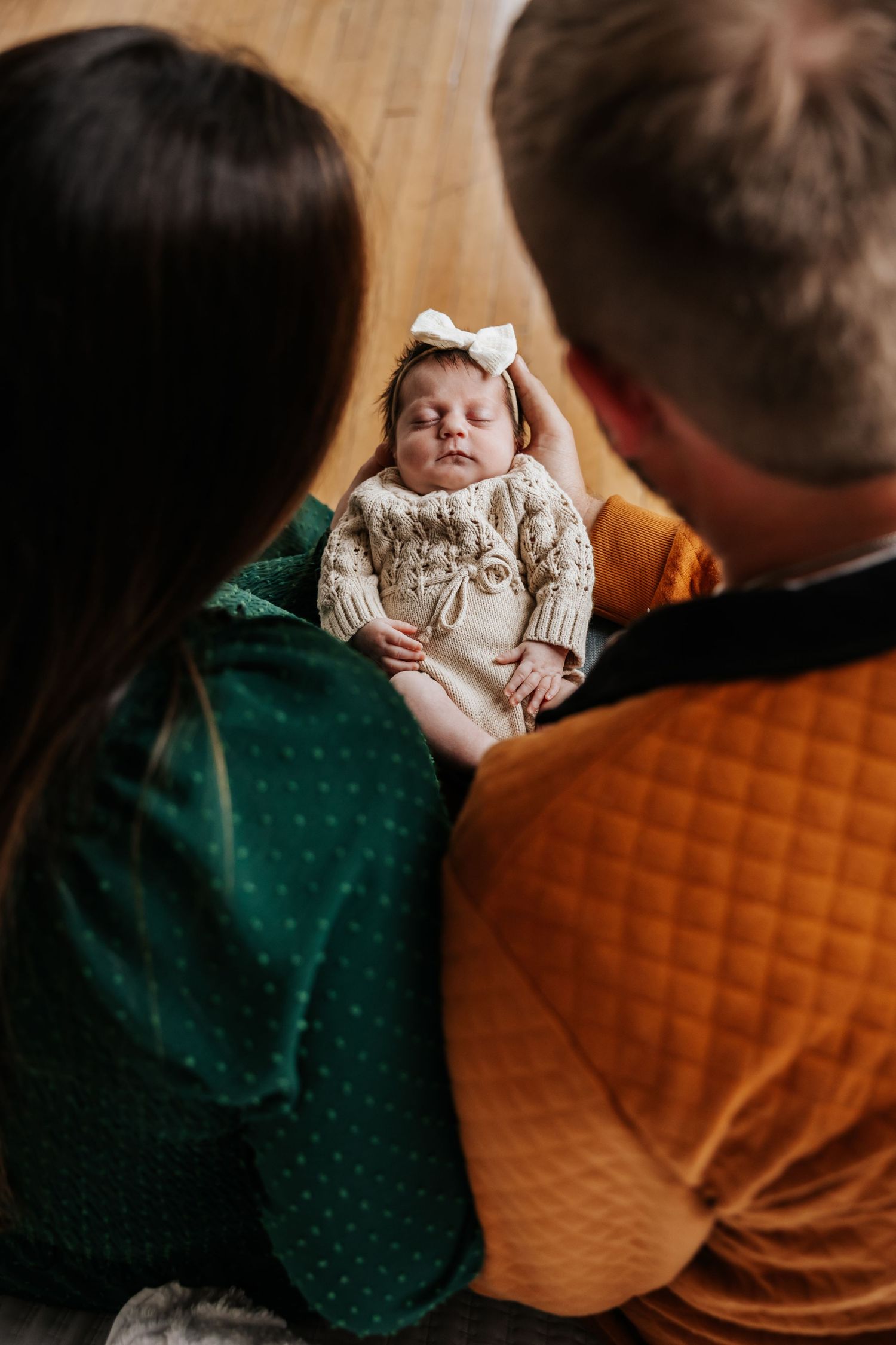A sleeping newborn girl in a knit outfit and white bow rests in her parents' arms in a tender overhead newborn portrait.