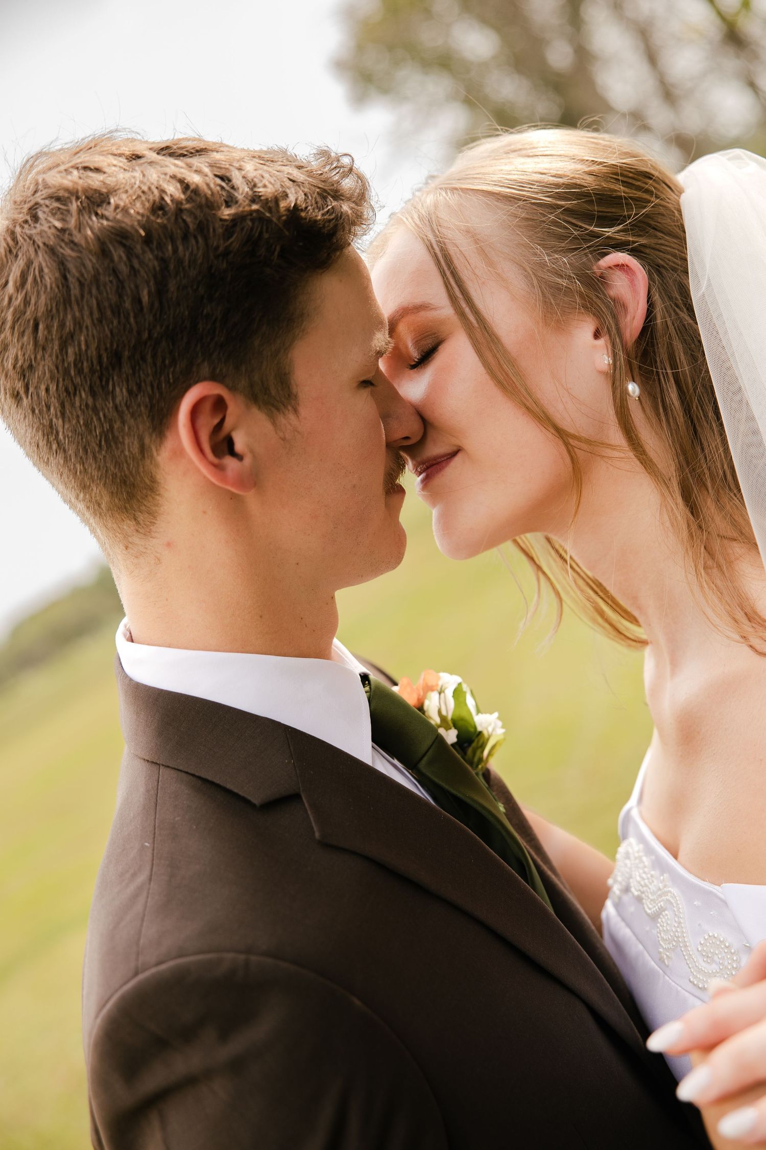 Close-up of a bride and groom touching noses in a romantic moment, with a floral boutonniere on his brown suit and a soft green backdrop.