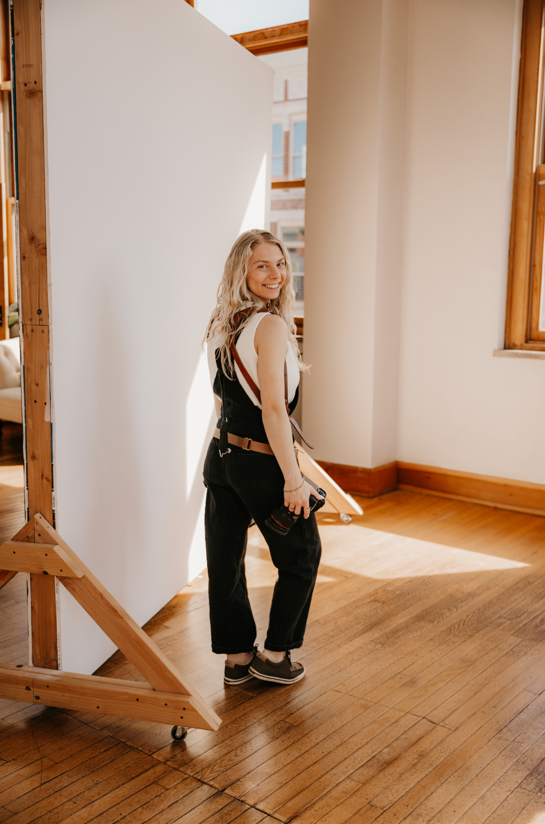 Smiling blonde photographer in black overalls glances back while carrying a camera in a sunlit wood-floor studio.