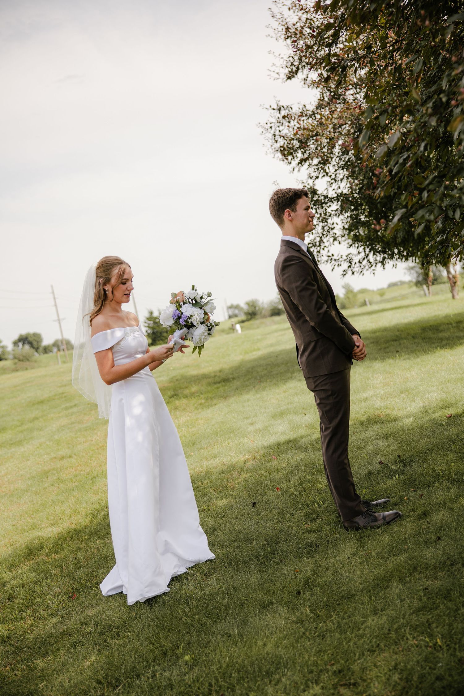 Bride in an off-shoulder white gown and veil holds a blue and white bouquet during a first look as the groom stands facing away on a lawn.